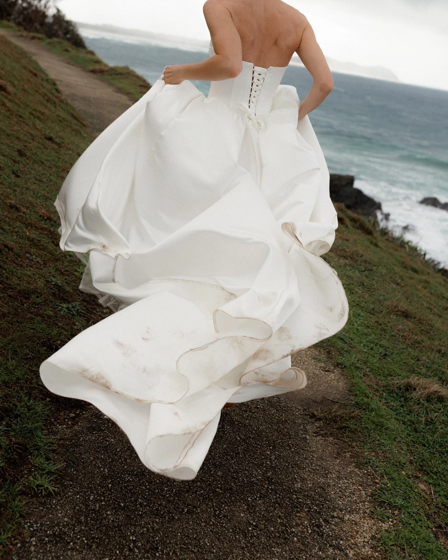 A person wearing a white wedding dress walking along a coastal path near the ocean with waves crashing onto rocks.