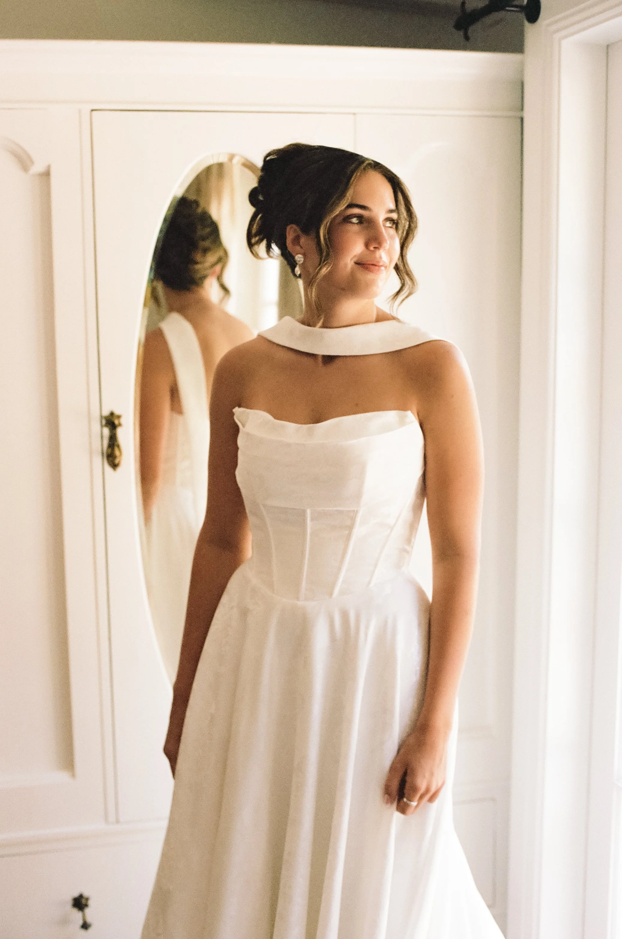 A woman in an elegant wedding dress standing in front of a mirror, with her reflection visible.