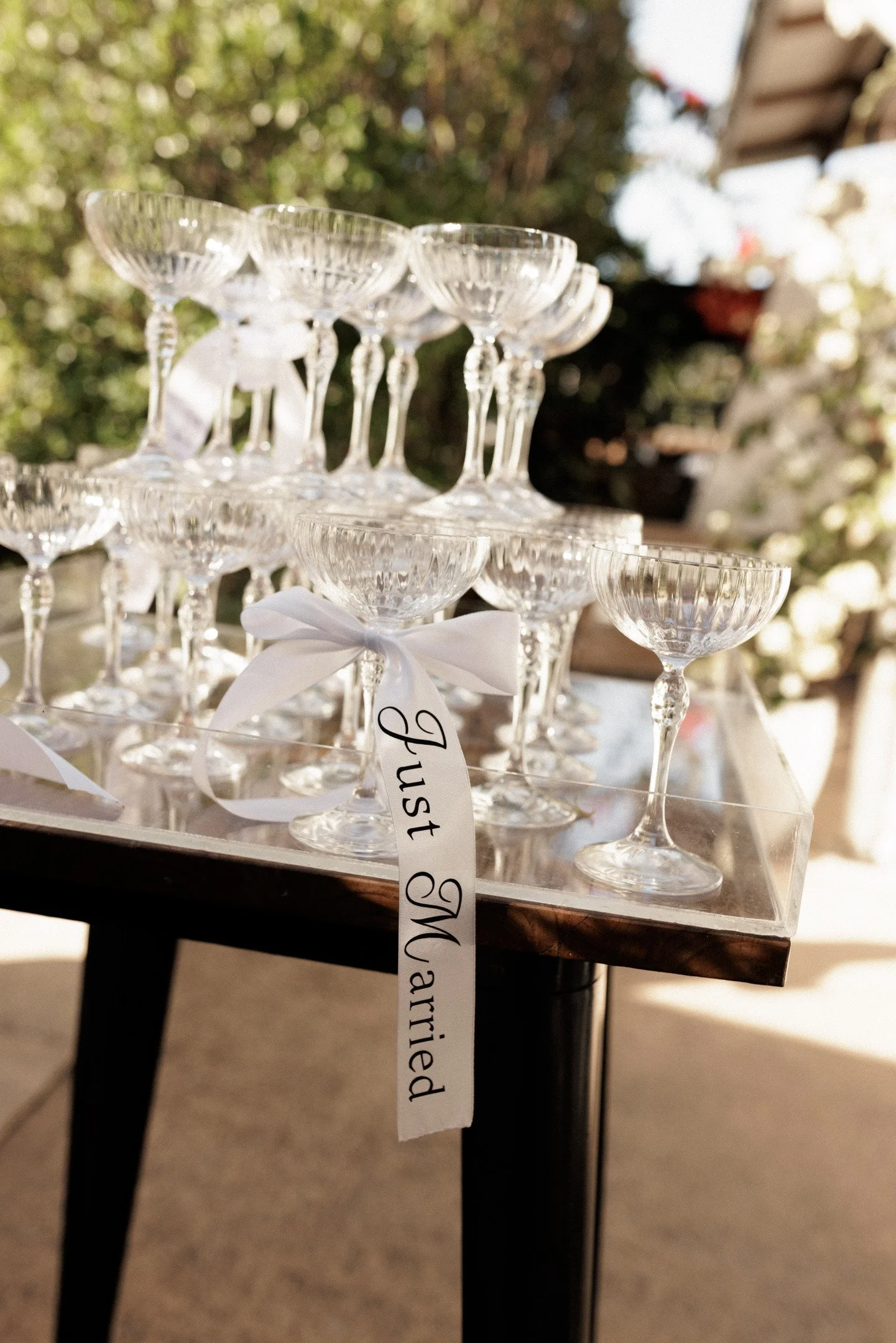 A display of empty glass dessert cups tied with a white ribbon, arranged on a table with a crystal-clear platform. In the background, blurred greenery and an outdoor setting are visible.