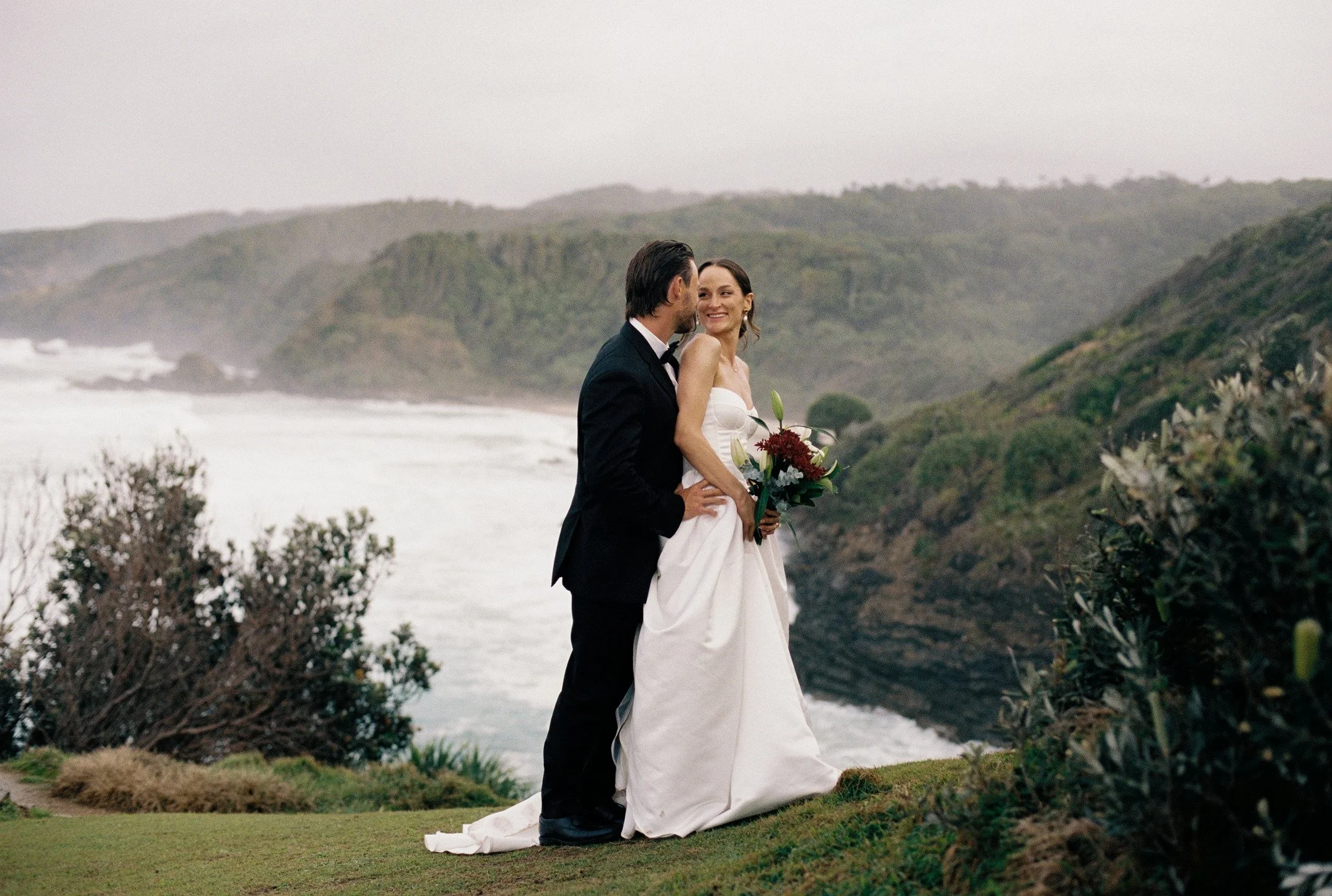 A bride and groom standing close together outdoors by the ocean, with lush green hills in the background, on a cloudy day. The bride is holding a bouquet and wearing a white wedding dress, while the groom is dressed in a black tuxedo.
