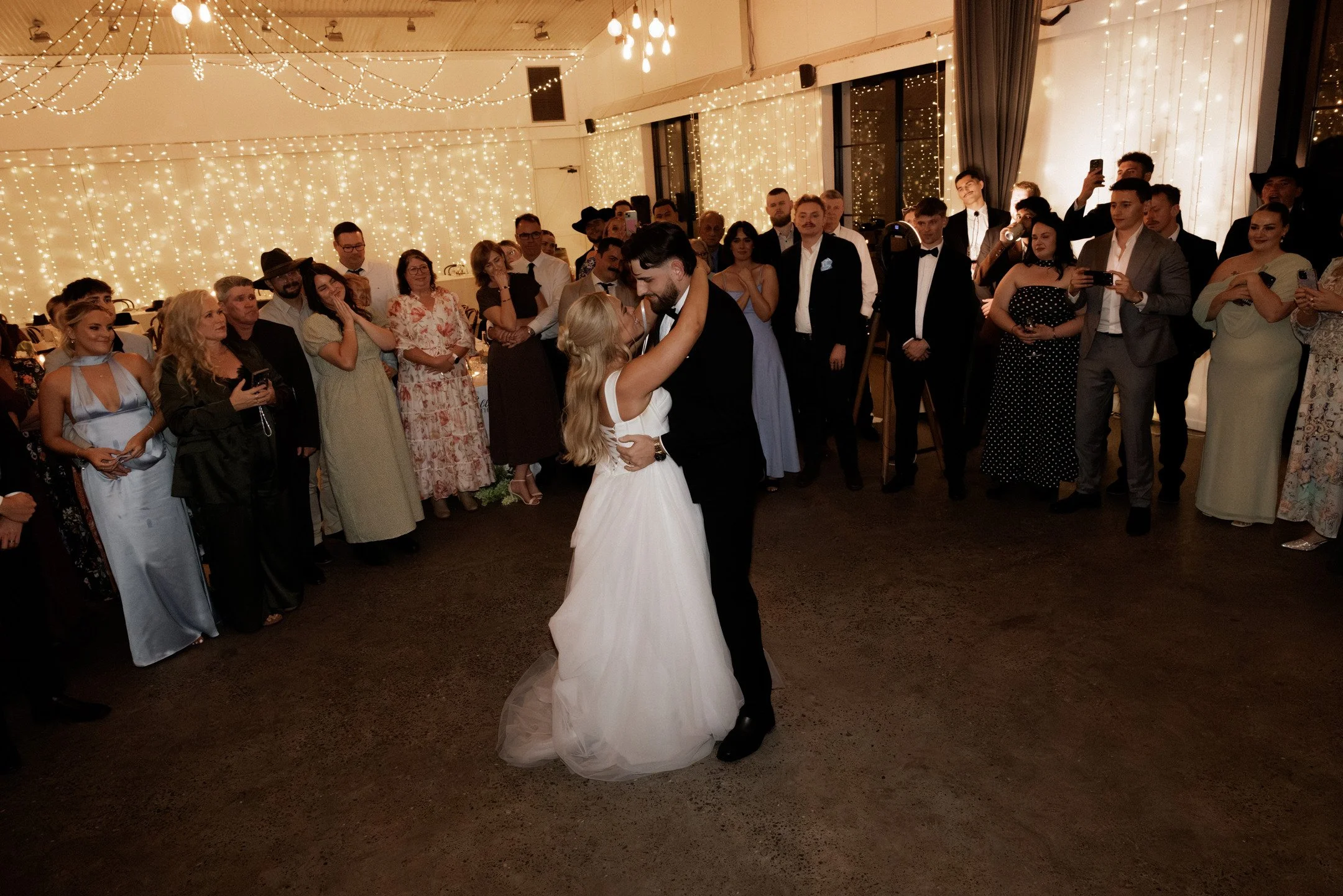 A bride and groom dancing at their wedding reception with guests watching and taking photos. The room is decorated with string lights and fairy lights.