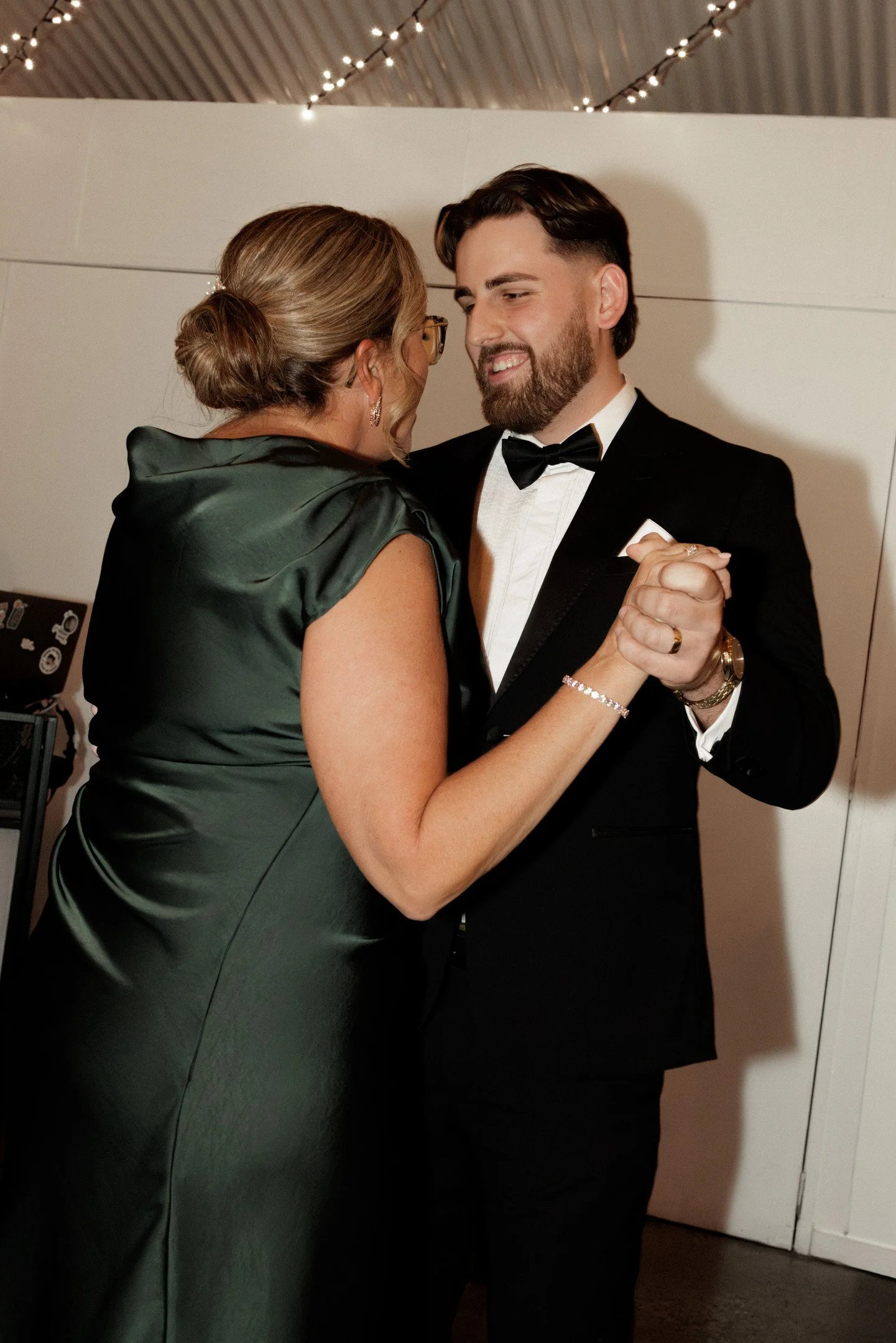 A woman and a man are dancing closely at a formal event. The woman has her hair styled in a bun, wears glasses, and a dark green satin dress. The man has dark hair, a beard, and is dressed in a black tuxedo with a white shirt and a bow tie. They are 