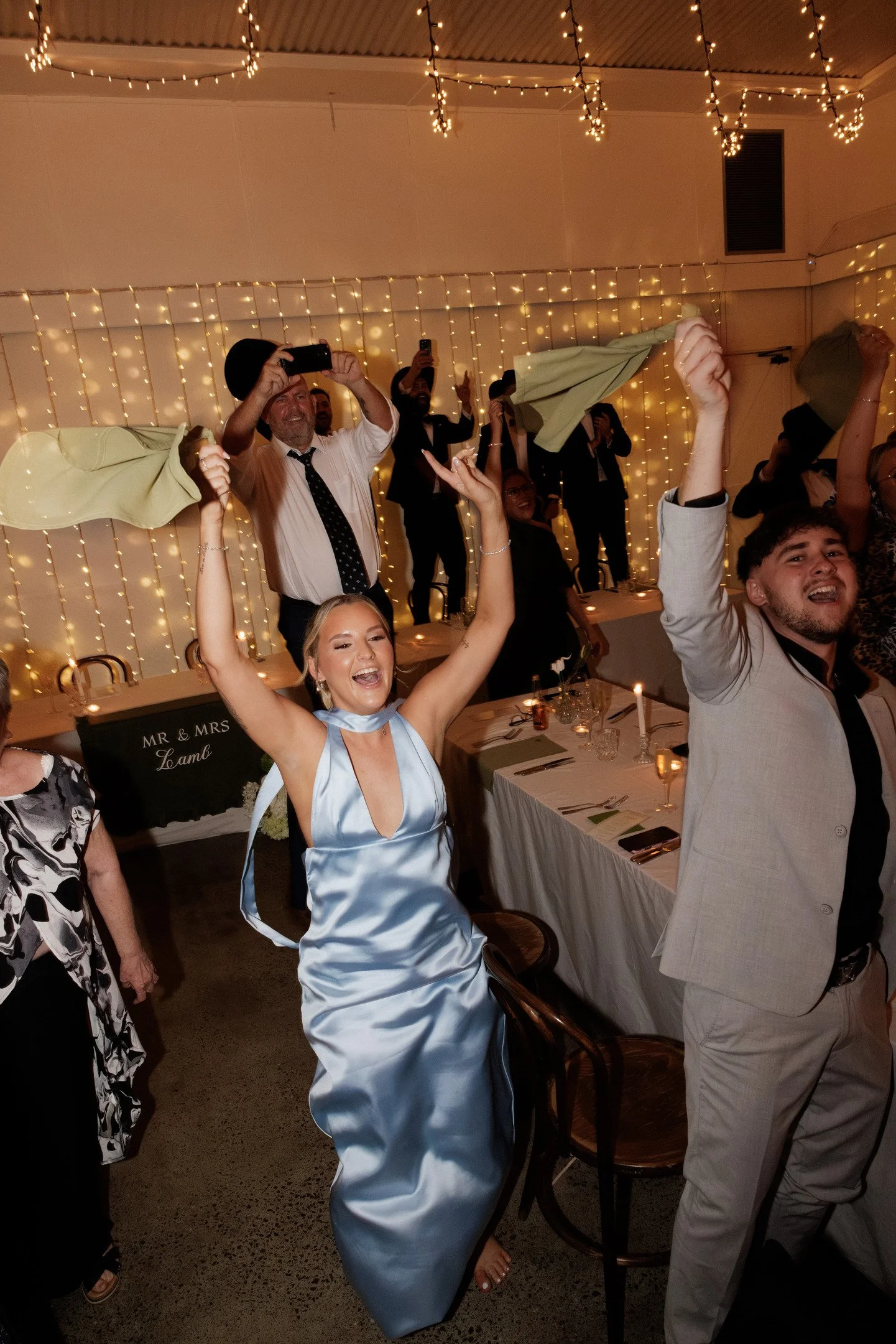 People celebrating at a party decorated with string lights, with some raising their arms and others taking photos, in a festive atmosphere.