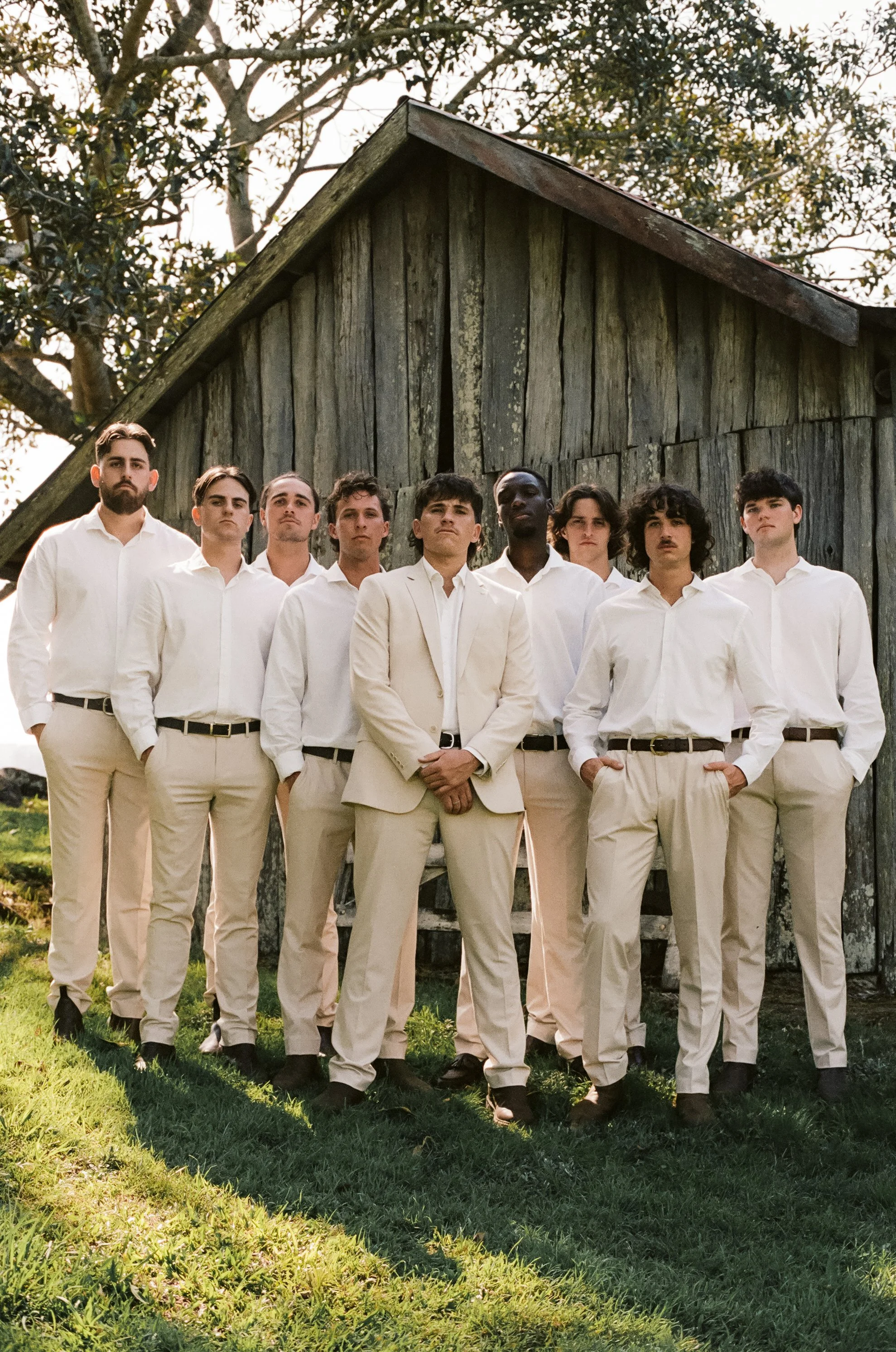 A group of nine young men in white and beige suits and shirts standing outside in front of a weathered wooden building with tall trees behind them.