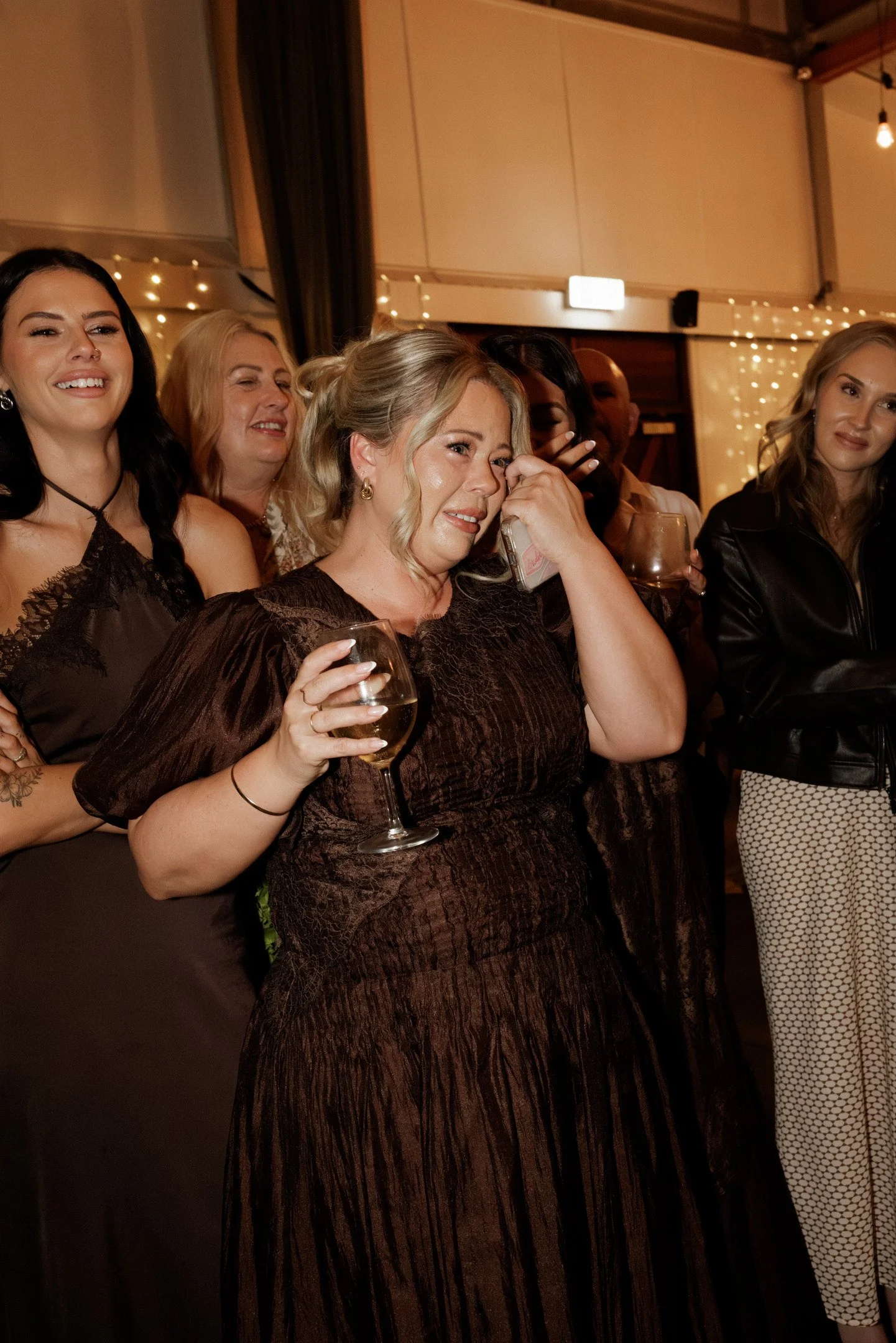 Group of women at a social event, some holding wine glasses, indoors with warm lighting and string lights in the background.