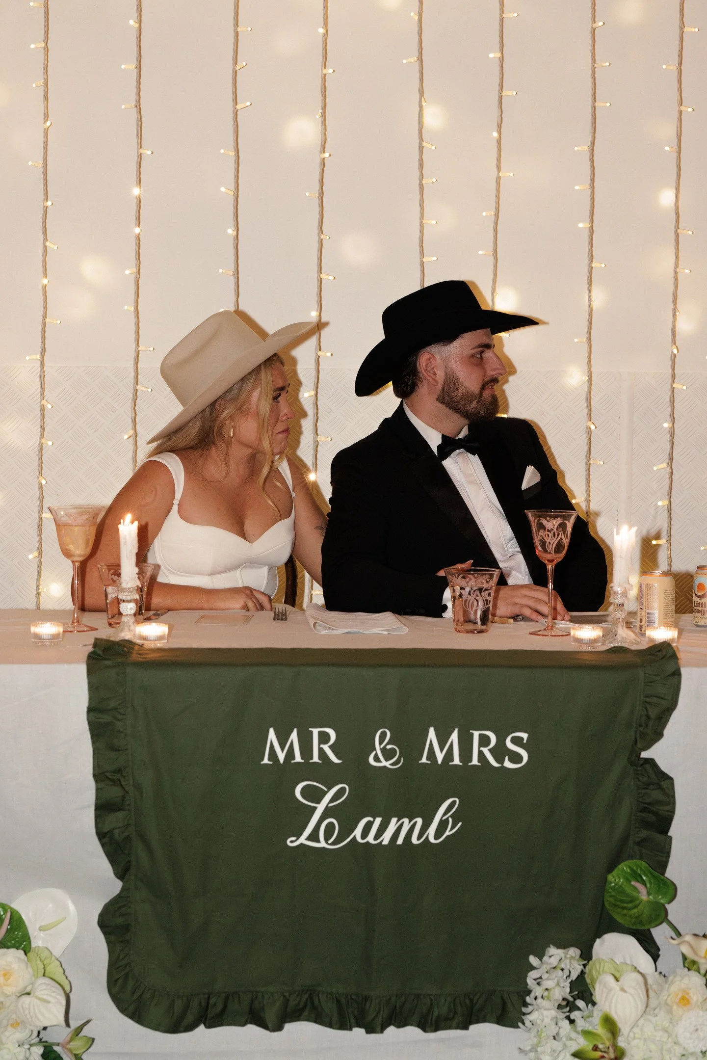 A wedding reception table with a green tablecloth that has white text saying 'MR & MRS Lamb.' Two people dressed in wedding attire are sitting behind the table, with a woman wearing a white dress and a beige cowboy hat on the left, and a man wearing 