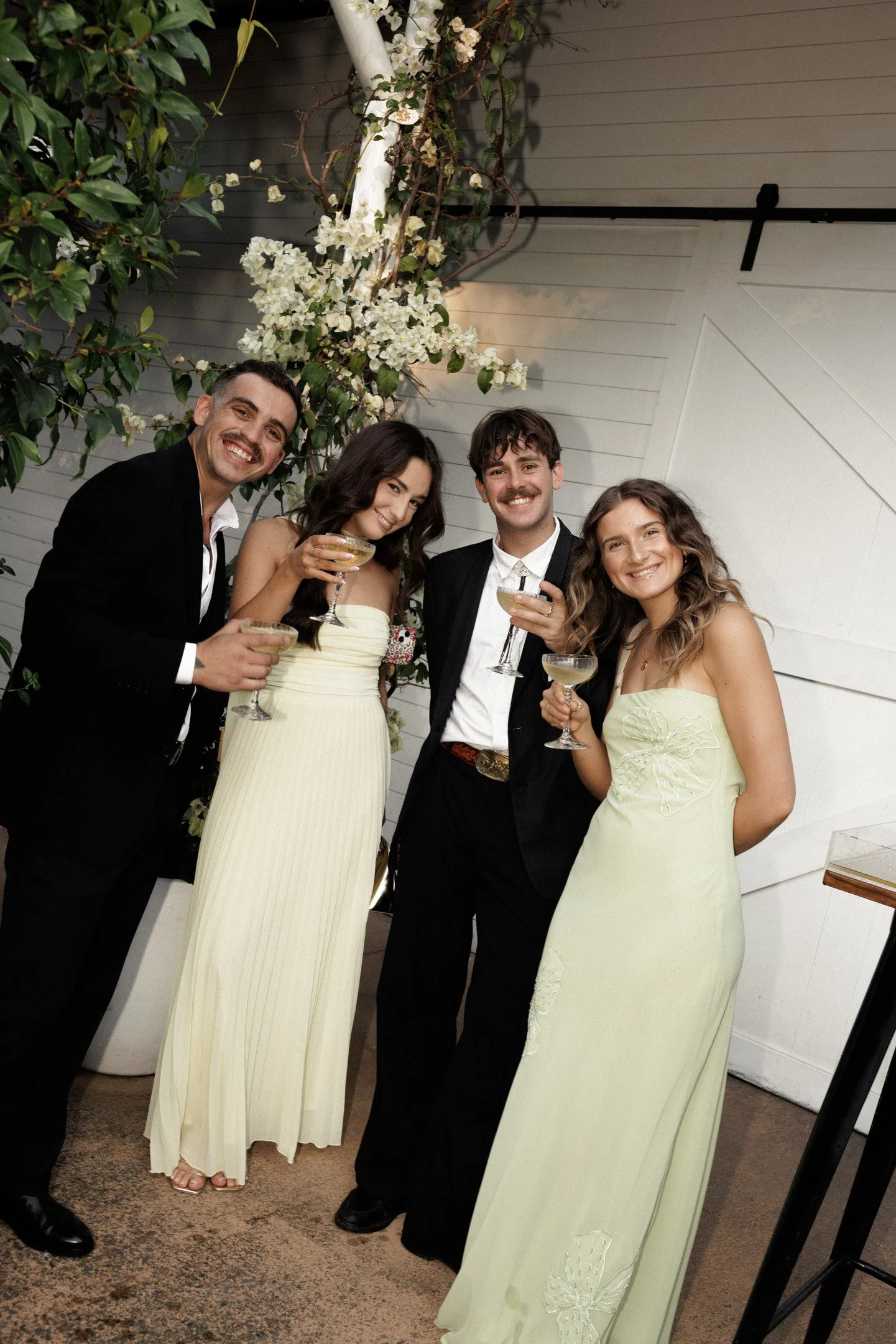 A group of four people dressed in formal attire enjoying drinks at an indoor event near a white wall with a decorative flower arrangement.