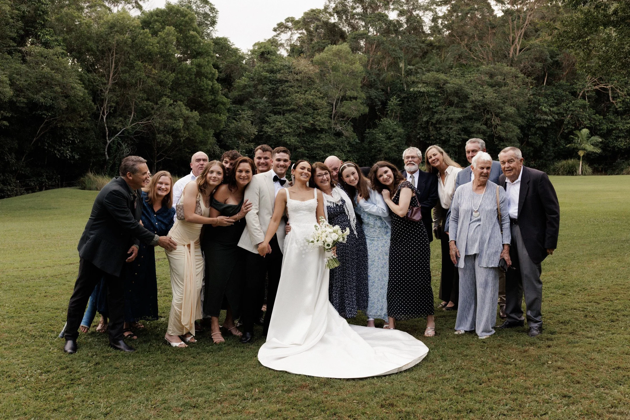 A group of people at a wedding, including the bride and groom, standing on a grassy area with a backdrop of trees.