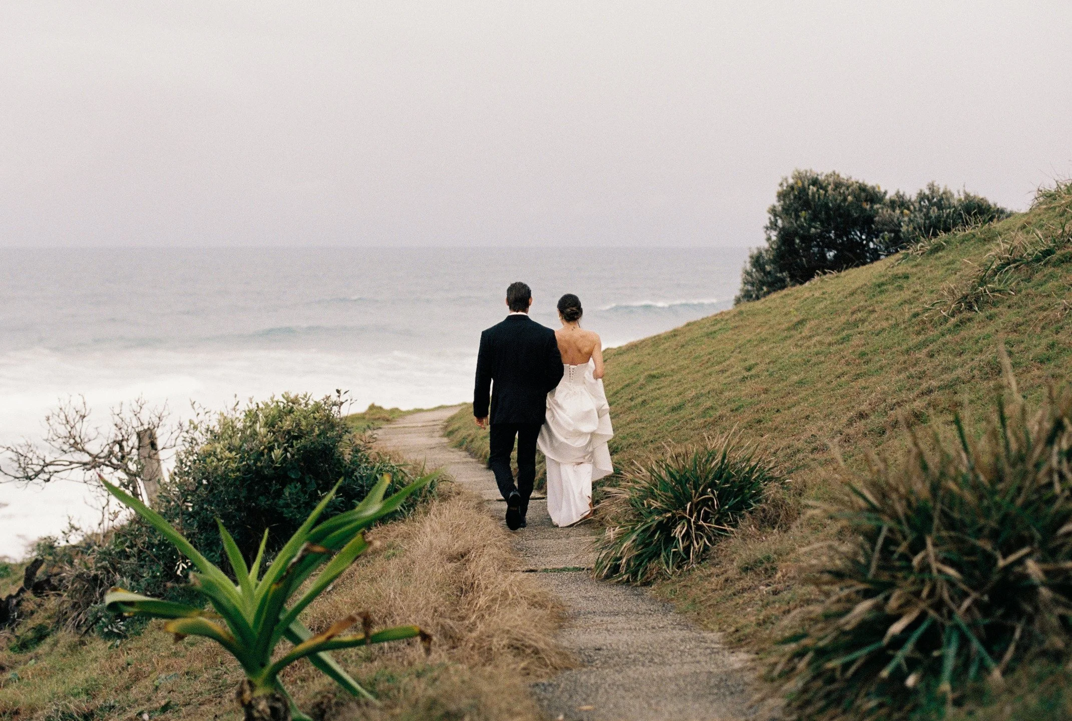 A couple walking along a pathway near the ocean, with the man in a black suit and the woman in a white dress, on a cloudy day with greenery surrounding the path.