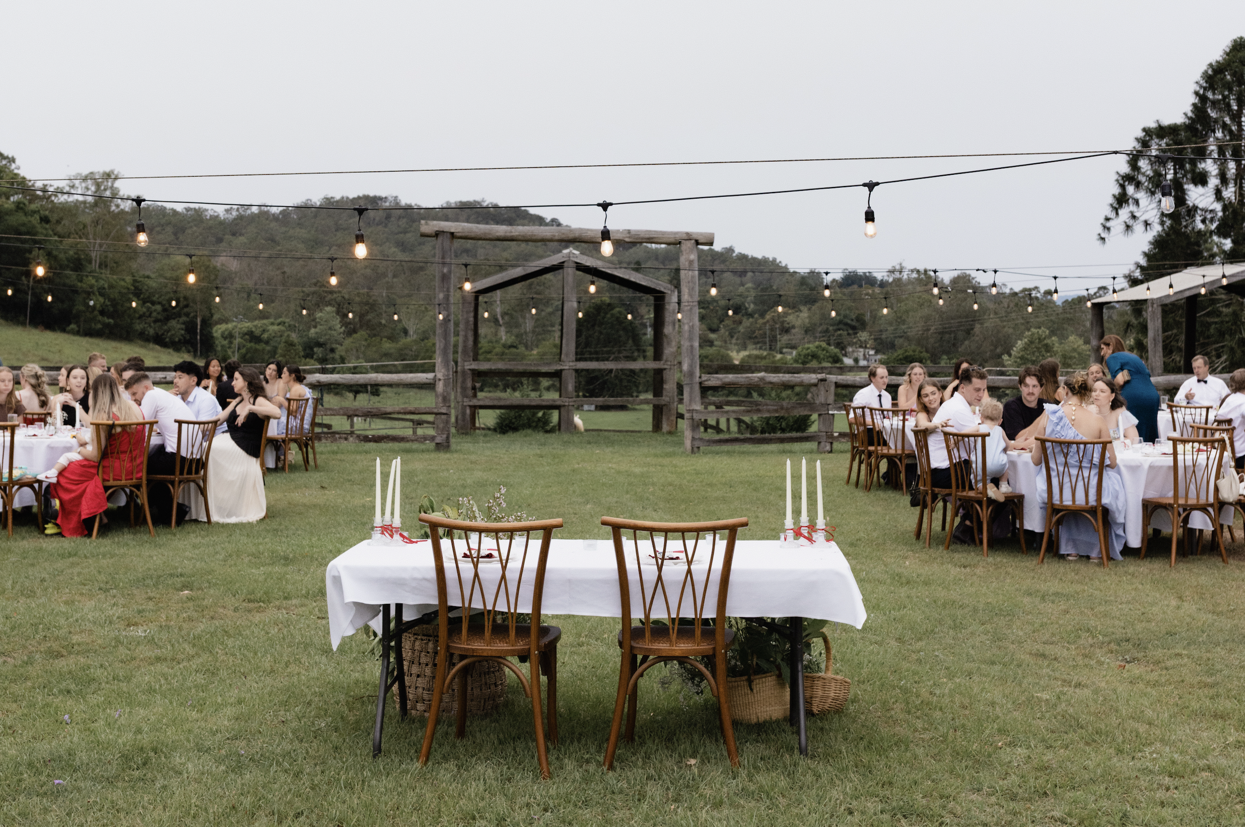Outdoor wedding reception with round tables covered in white tablecloths, decorated with candles and flowers, set on a grassy field under string lights, with a wooden gate and green hills in the background.
