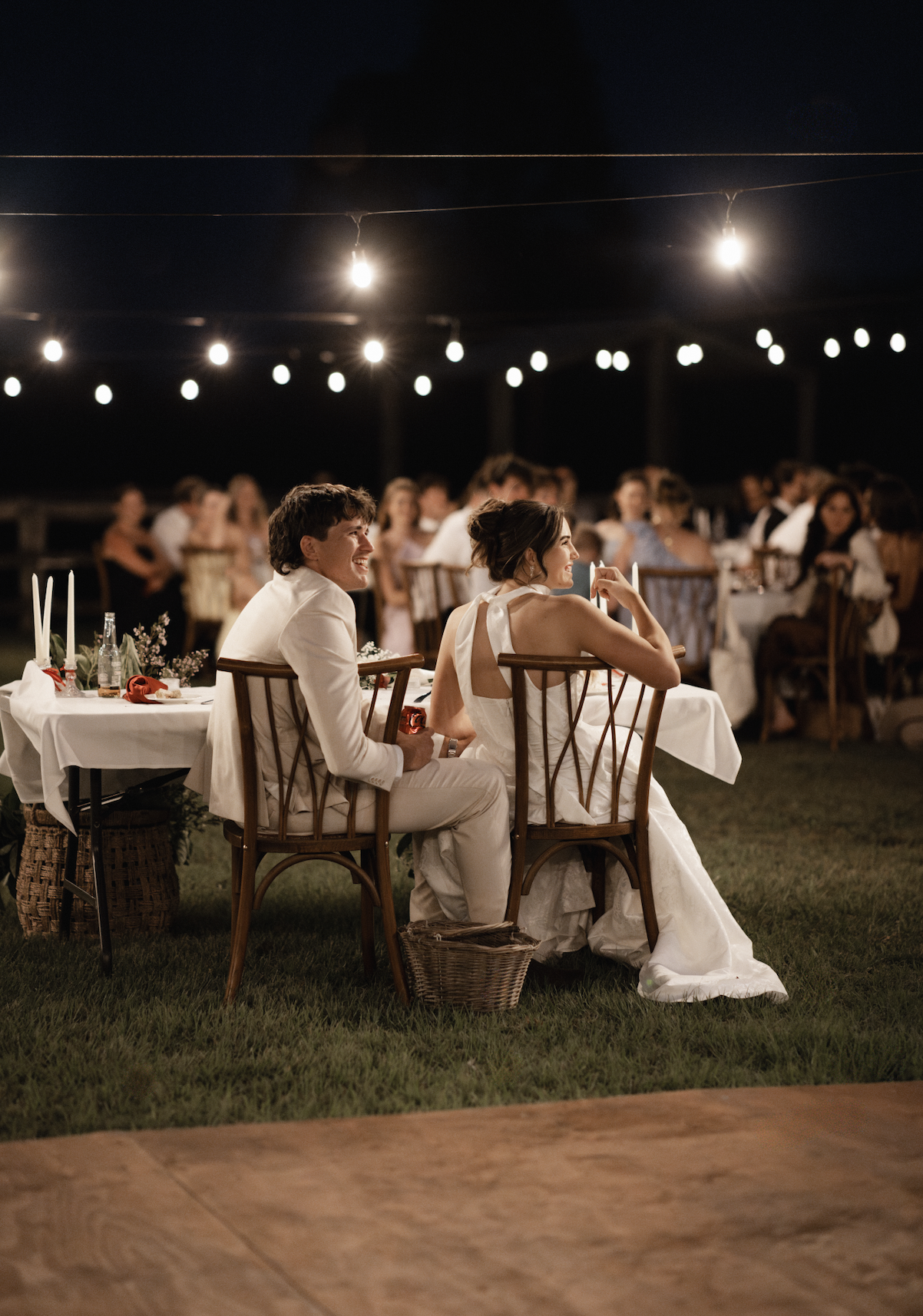 A wedding reception at night with a bride and groom sitting at a table, surrounded by guests, under string lights.