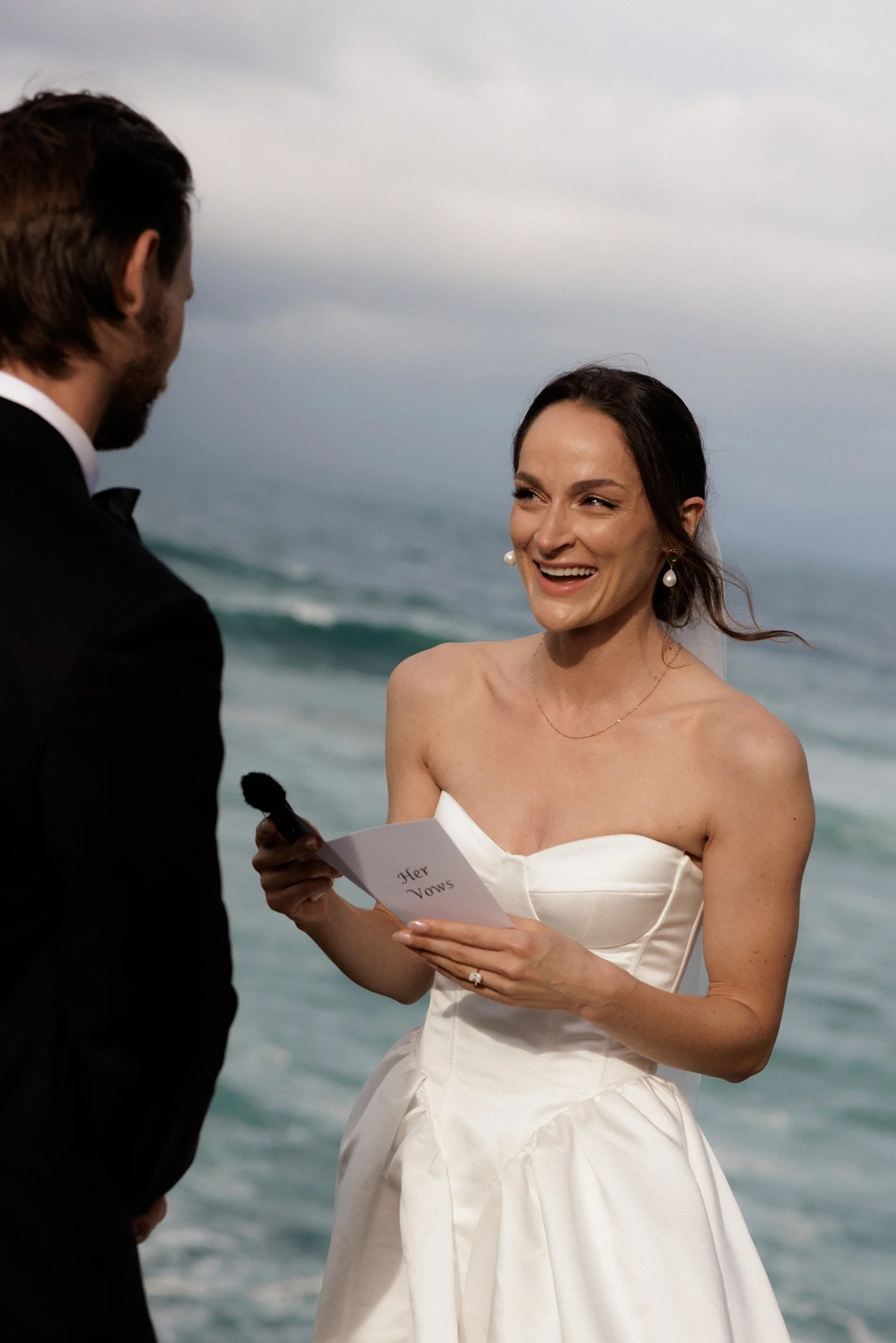 A woman in a strapless white wedding dress smiling and holding a piece of paper with 'Her Vows' written on it, standing by the ocean, during her wedding ceremony.