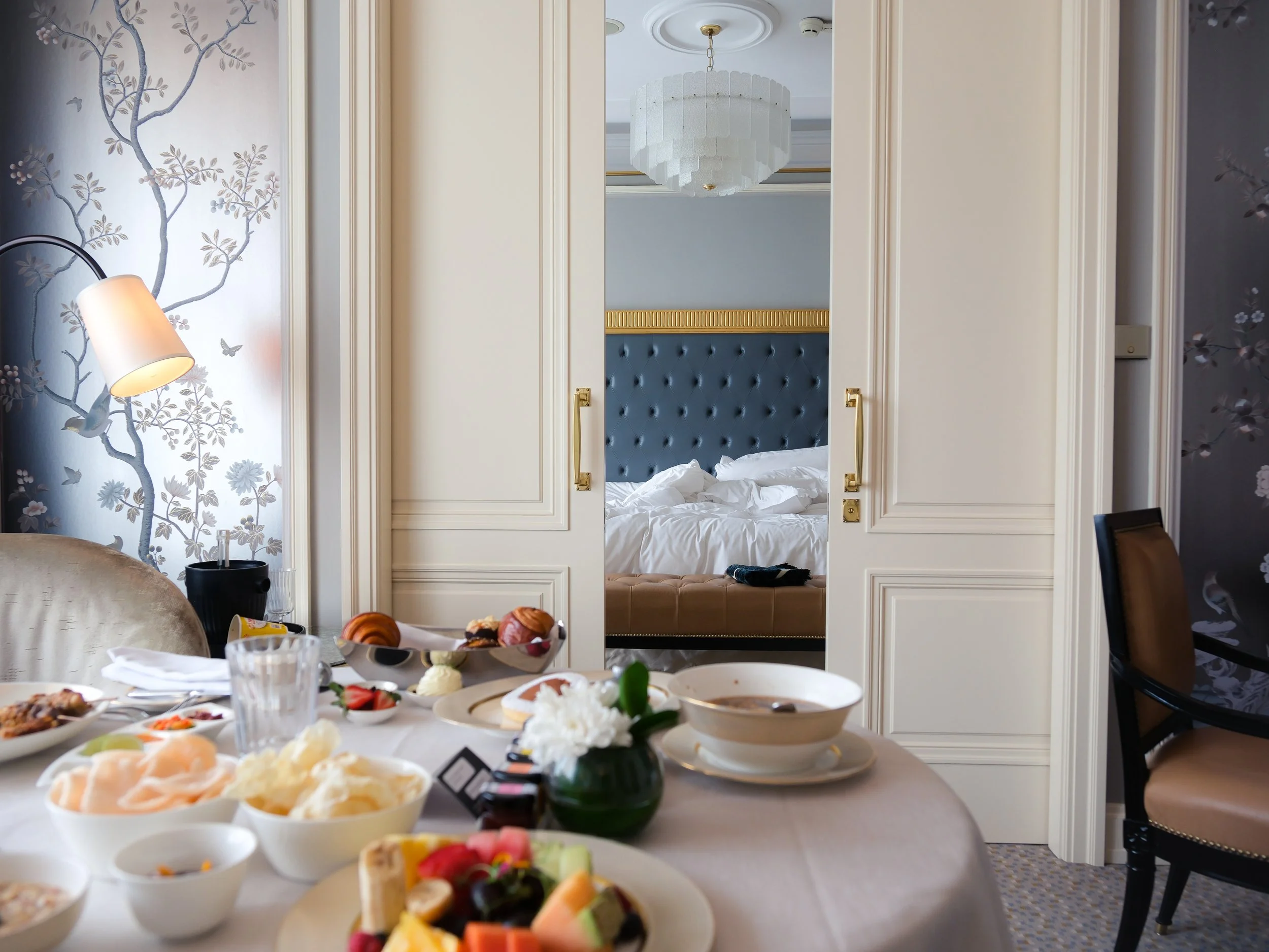 A dining table filled with various dishes including bowls of fruit, desserts, and snacks, with a bedroom visible through an open door in the background.
