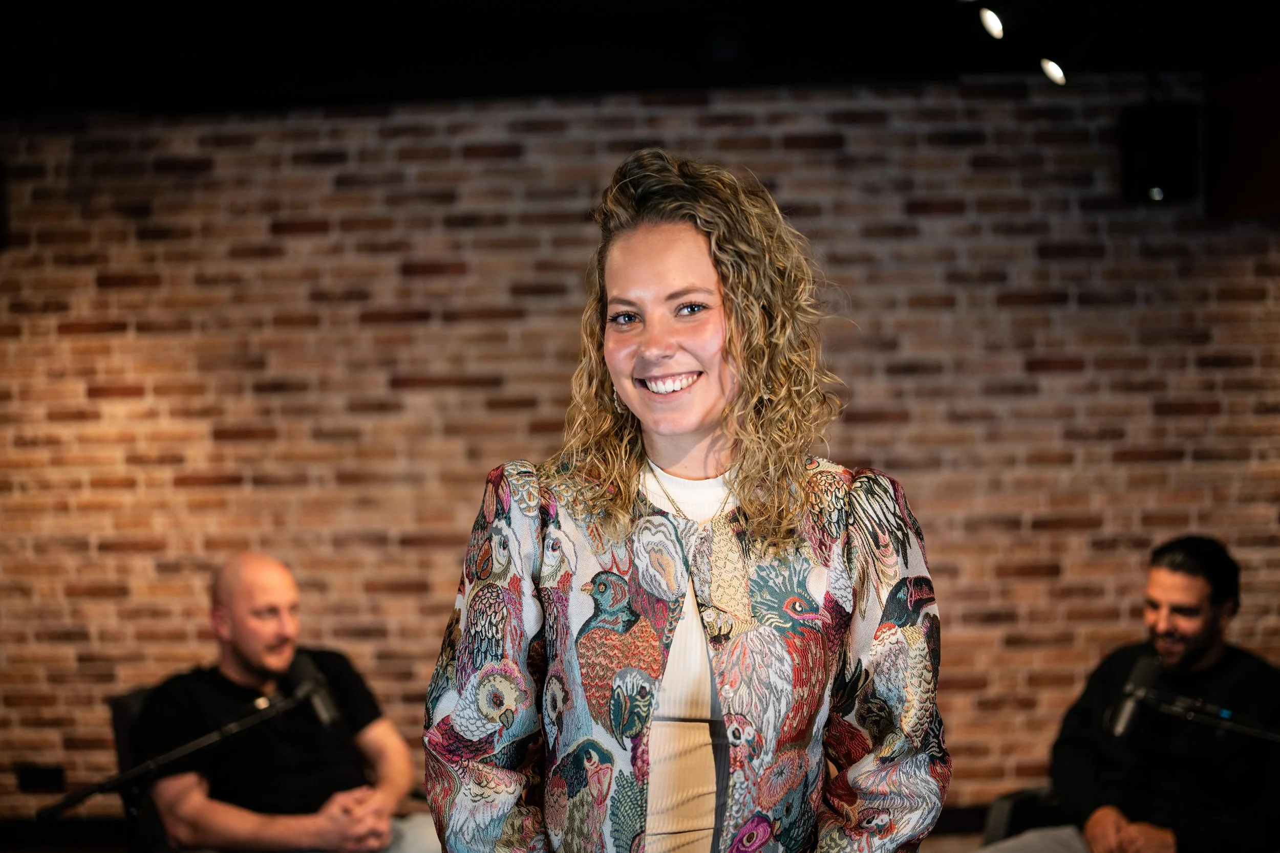 twee mannen in gesprek in de achtergrond terwijl de podcast manager van podcast studio westland lachend naar de camera kijkt