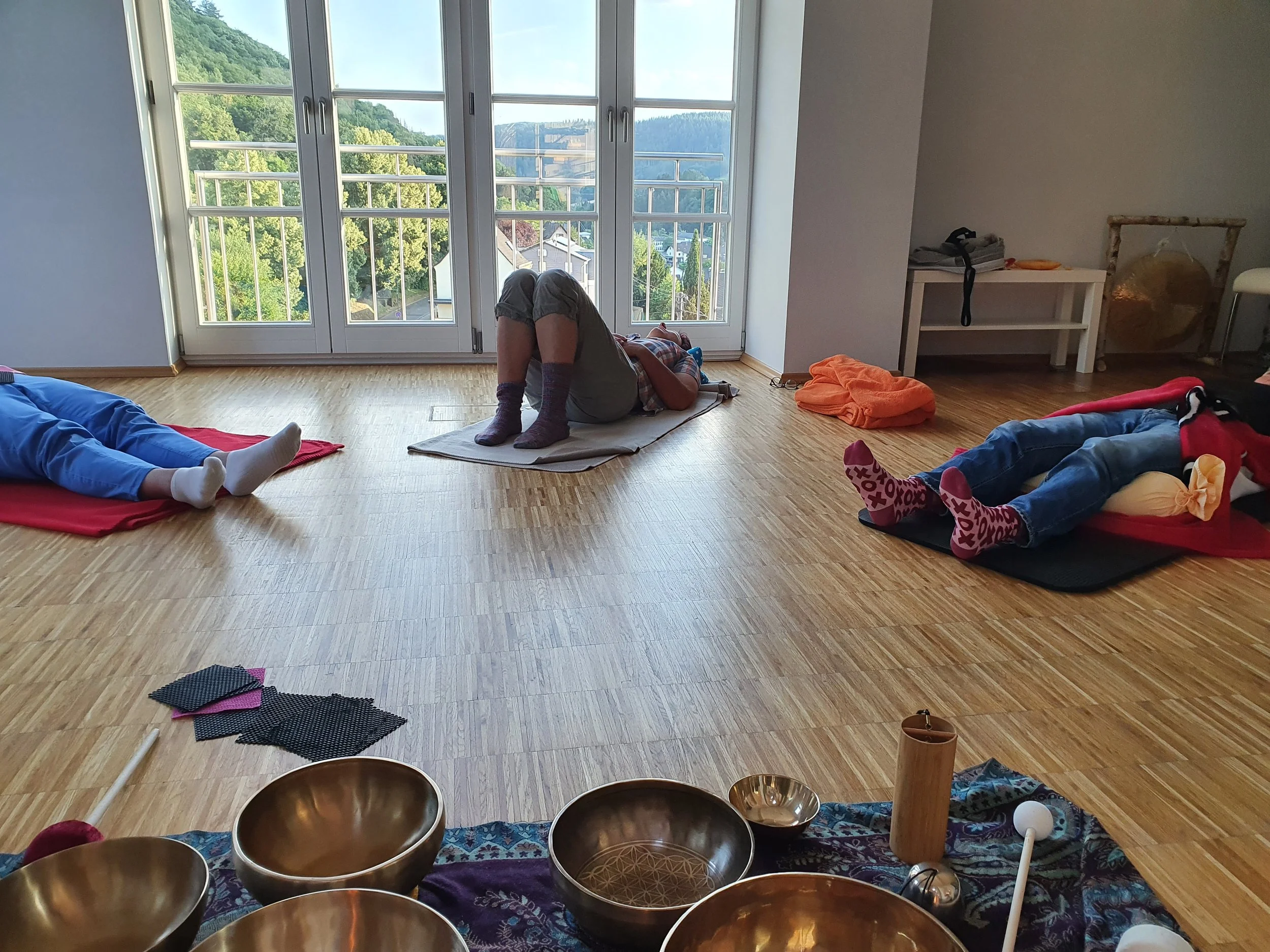 Menschen bei einer Klangschalen-Meditation in einem hellen Raum mit Holzboden und großen Fenstern, die eine grüne Landschaft zeigen.