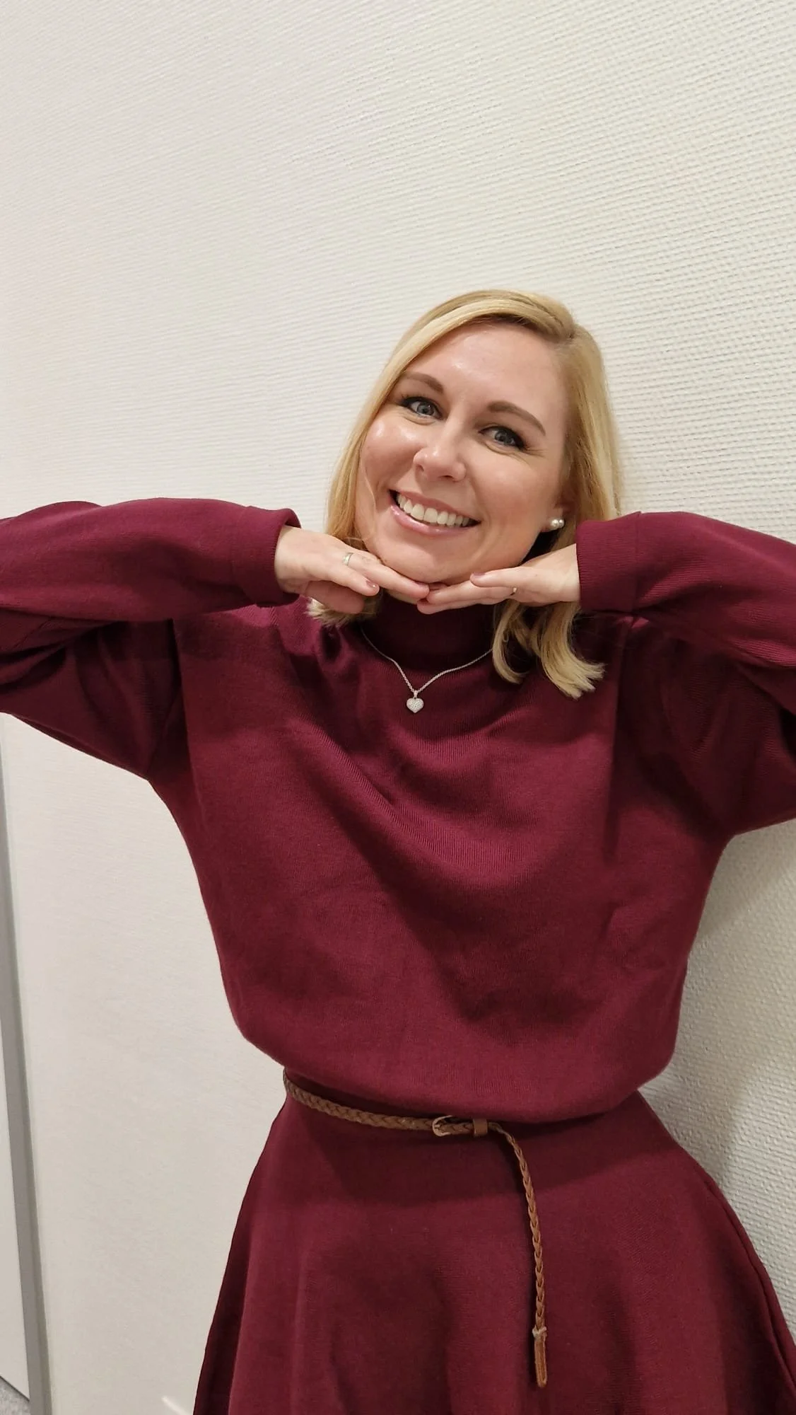 Eine lachende blonde Frau in rot-rosa Kleidung, die die Hände unter dem Kinn hält, stehend vor einer weißen Wand.