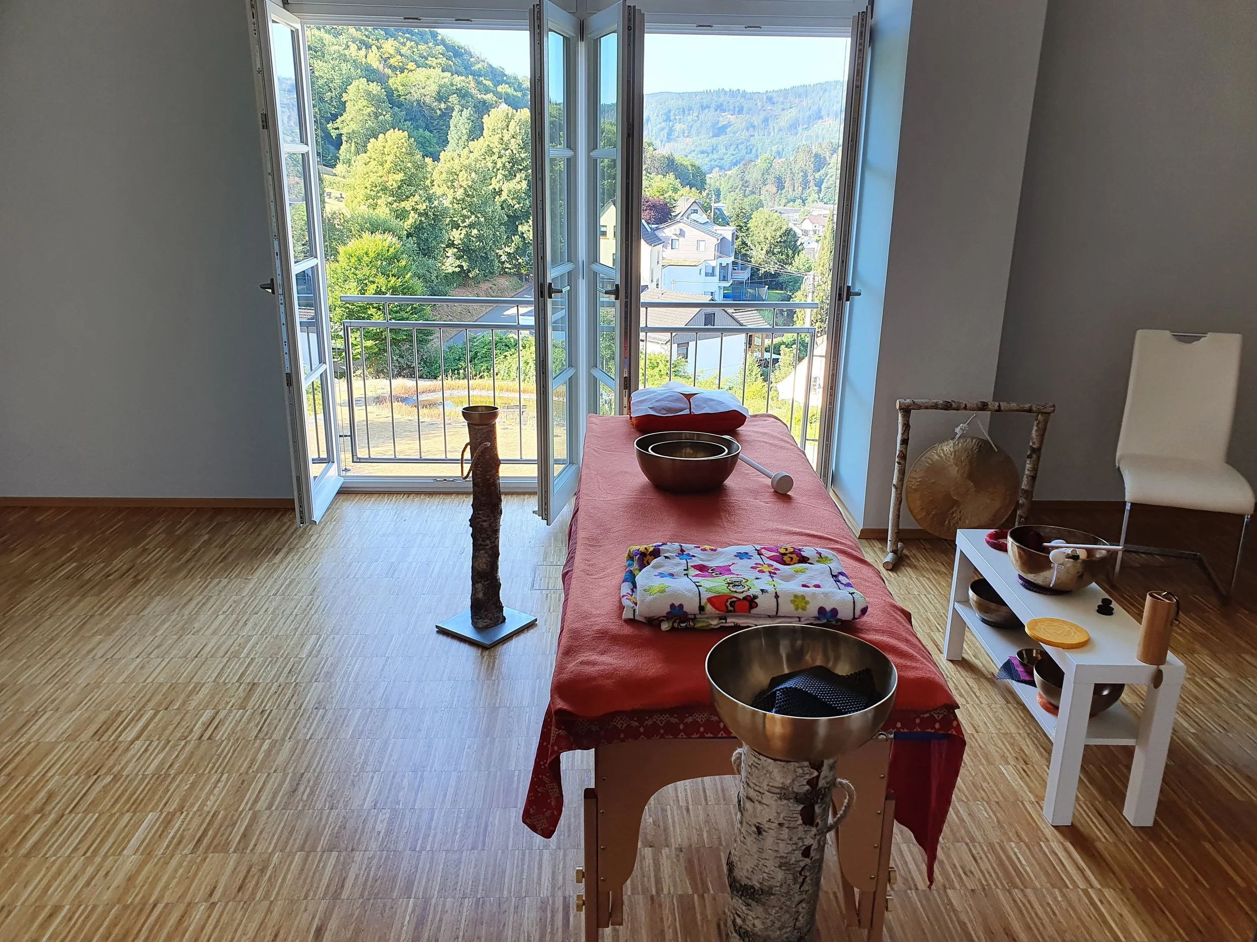 Innenraum eines Zimmers mit offenem Balkon, einem Behandlungs- oder Massagebett, einem Massage-Gong, einer Klangschale im Rahmen, einer weißen kleinen Tisch mit weiteren Klangschalen, einem weißen Stuhl, und einer großen Fensterfront mit Blick auf eine grüne Landschaft mit Häusern und Hügeln.