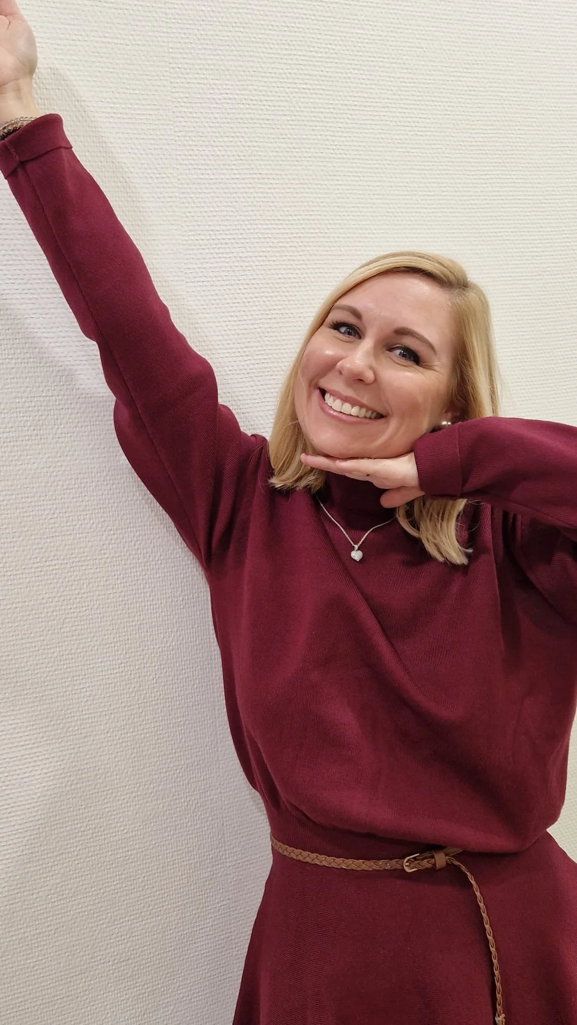 Lächelnde Frau in rotem Pullover, die die rechte Hand hochhebt und die linke Hand unter das Kinn legt, vor einer weißen Wand.