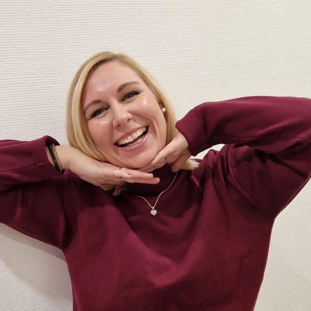 Lächelnde Frau mit blondem Haar trägt ein bordeauxrotes Sweatshirt, posiert vor weißer Wand, hält Kopf mit Händen.