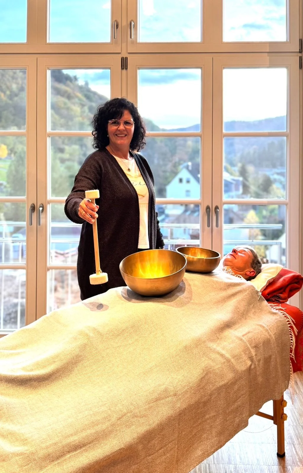 Eine Frau hält einen Gong-Schlägel vor einem Gong. Vor ihr liegt eine Massagebank mit einem Menschen, der entspannt liegt. Der Raum hat große Fenster mit Blick auf die Landschaft draußen.