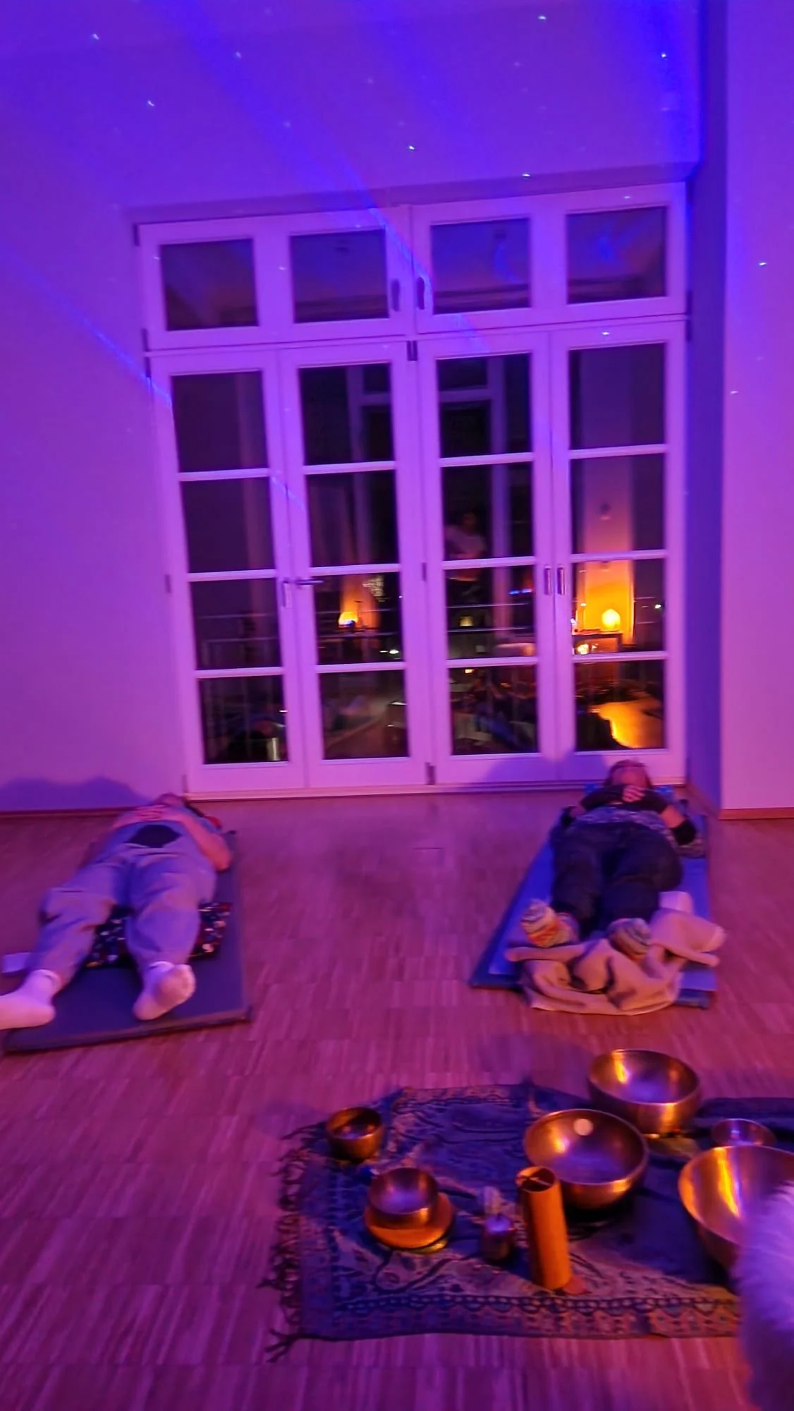 Zwei Kinder liegen auf Yogamatten in einem Raum mit großen Fenstern, die in den Abendhimmel blicken, beleuchtet mit buntem Licht, mit Klangschalen auf einem Teppich