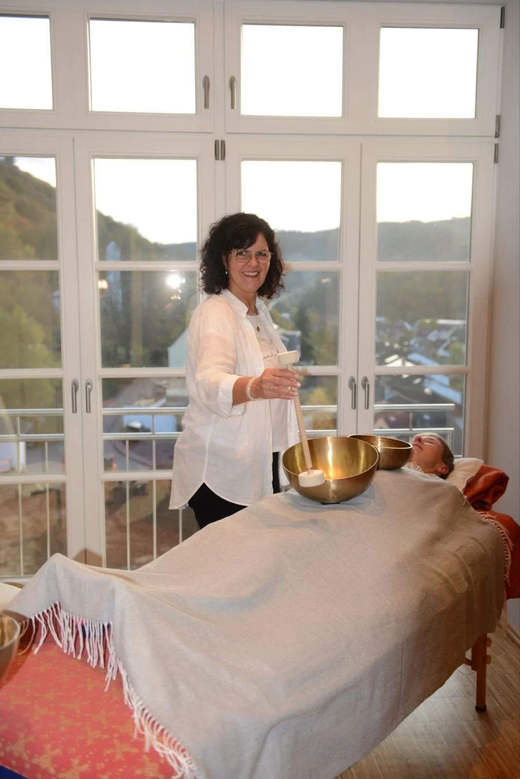 Eine Frau macht eine Klangtherapie mit einer Hartholzklangschale auf einer Massagebank, während eine andere Person entspannt liegt.