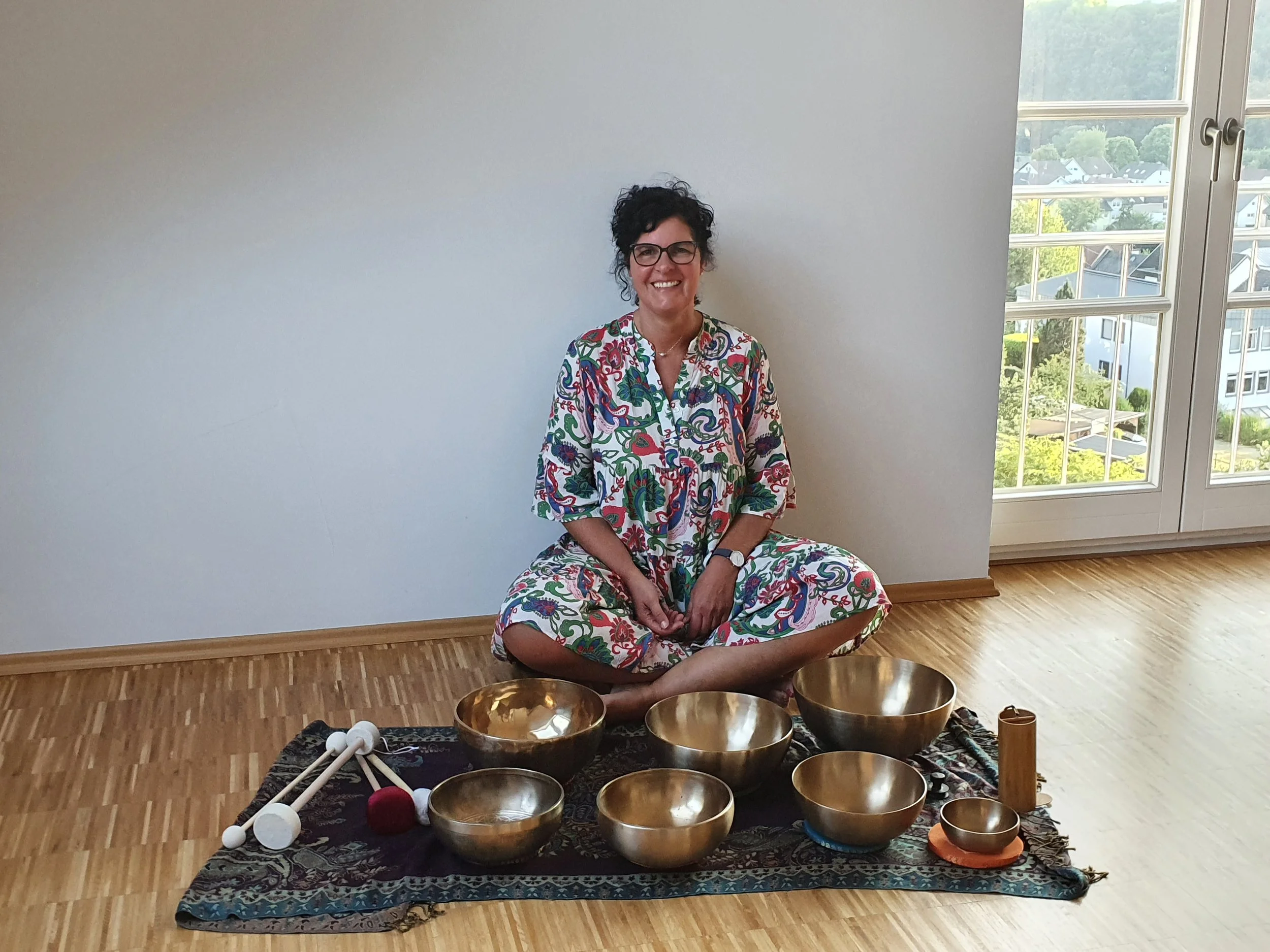 Frau sitzt im Schneidersitz auf Holzparkett, umgeben von Klangschalen und einem Meditationstool, vor einem weißen Wand und großen Fenstern mit Blick auf eine grüne Landschaft.