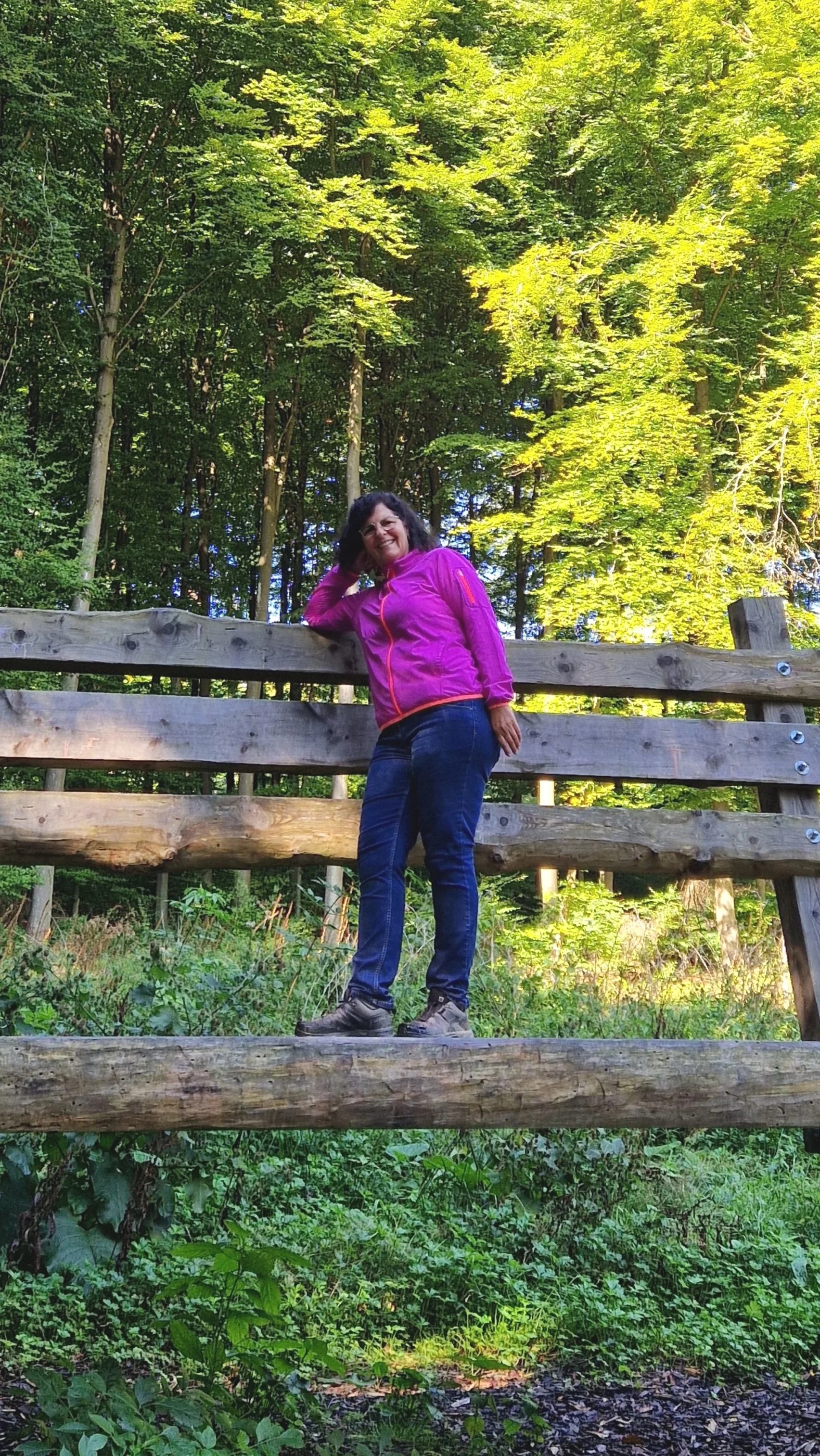 Frau in pinker Jacke steht auf einer Holzbrücke im Wald, umgeben von grünen Bäumen.