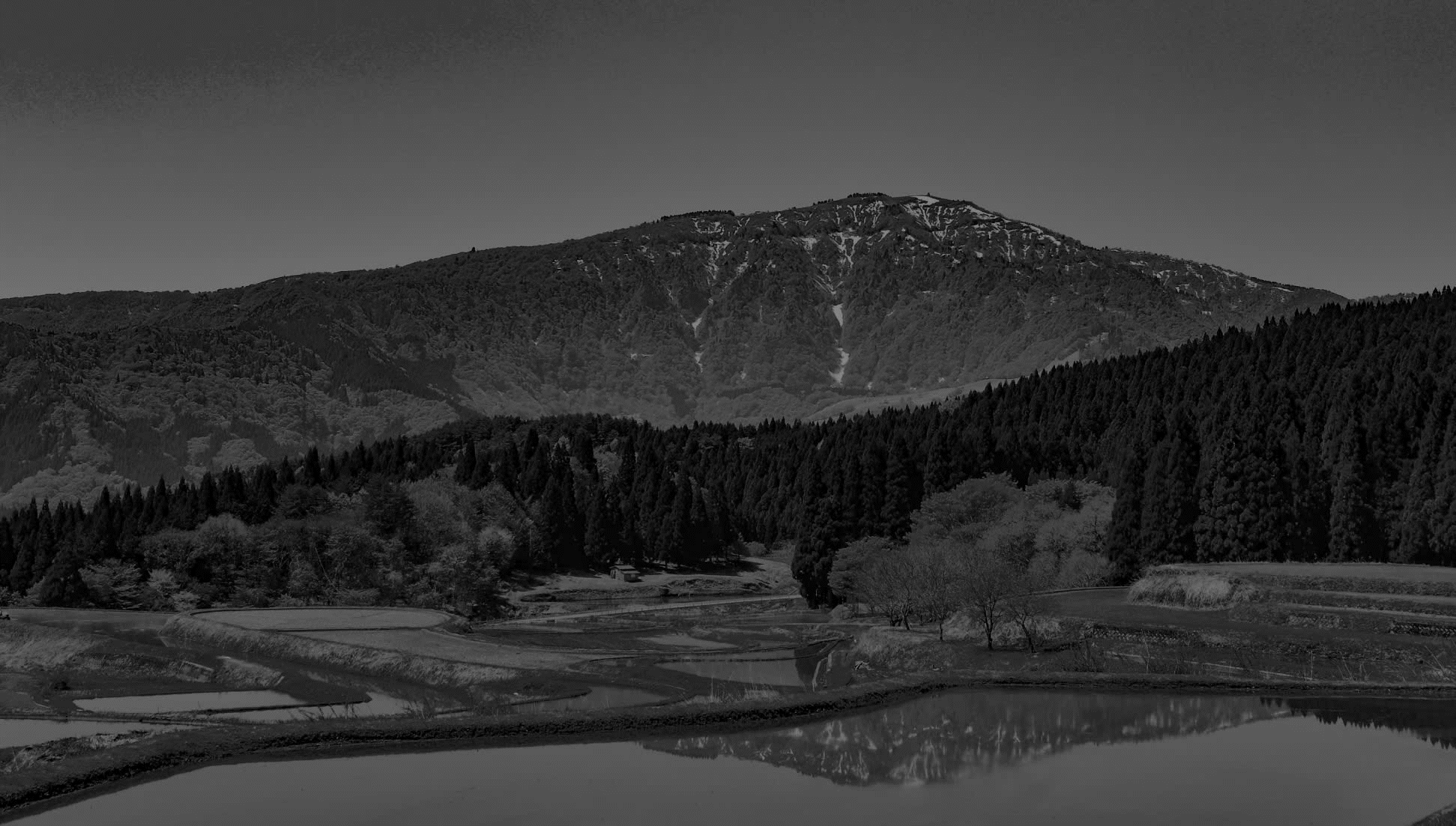 Scenic landscape of a mountain with forested slopes, terraced fields in the foreground, and a small body of water reflecting the scene.