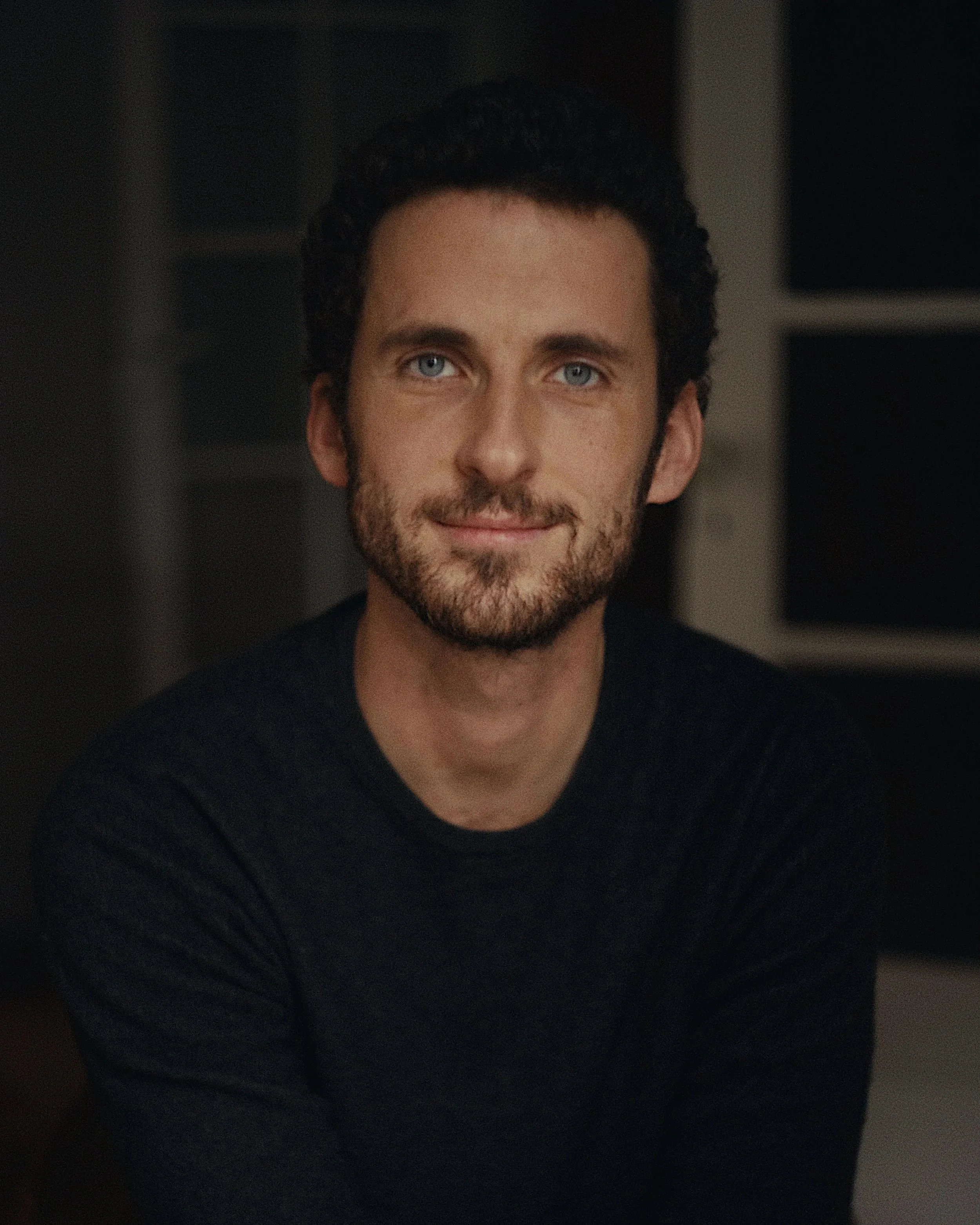 Un homme avec des cheveux bouclés et une barbe, portant un t-shirt noir, souriant légèrement, dans un environnement intérieur