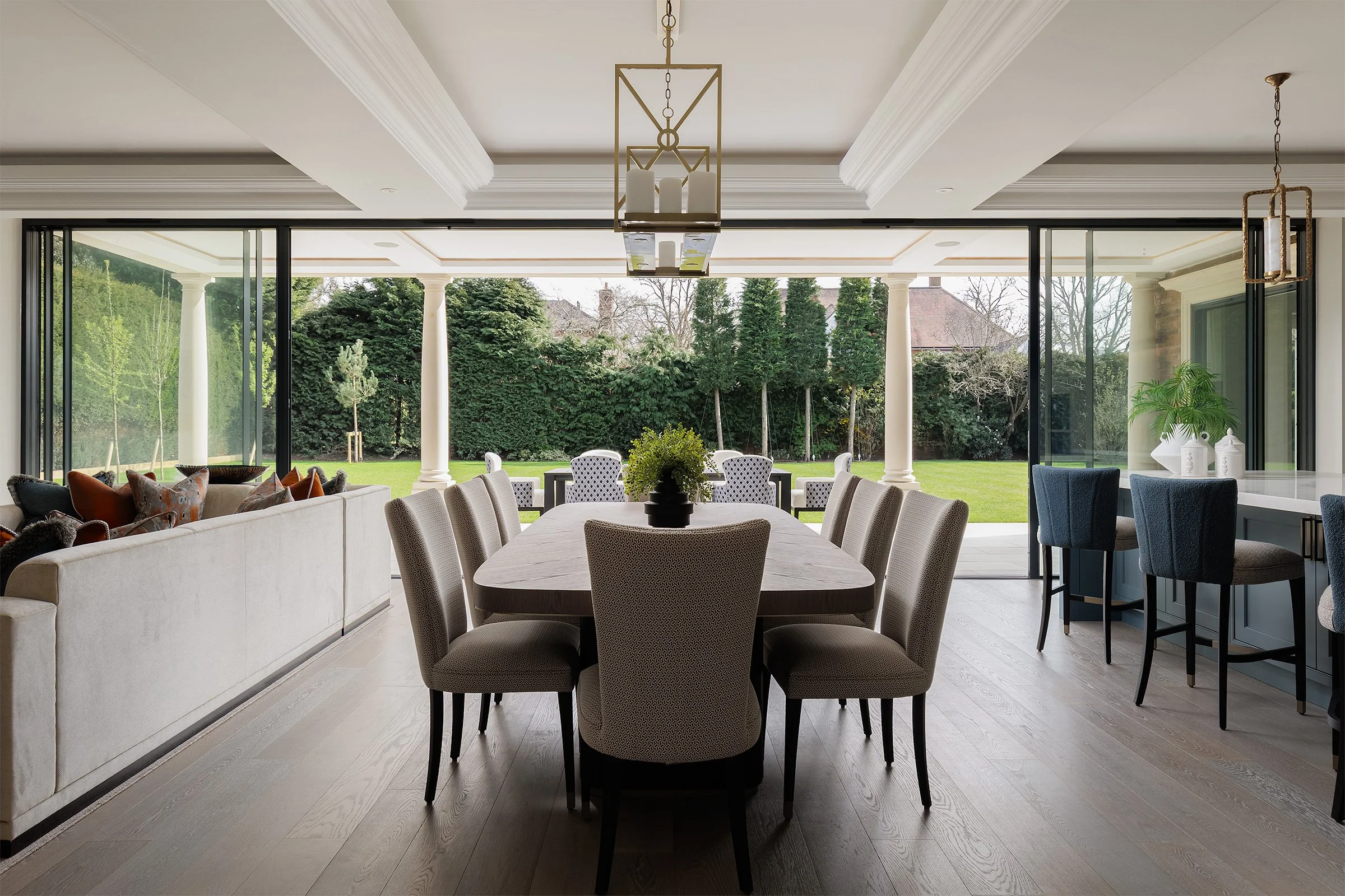Elegant dining room with a large table and upholstered chairs, set in an open space with floor-to-ceiling glass doors leading to a manicured garden. Features modern chandeliers, a cozy sitting area, and a breakfast bar.