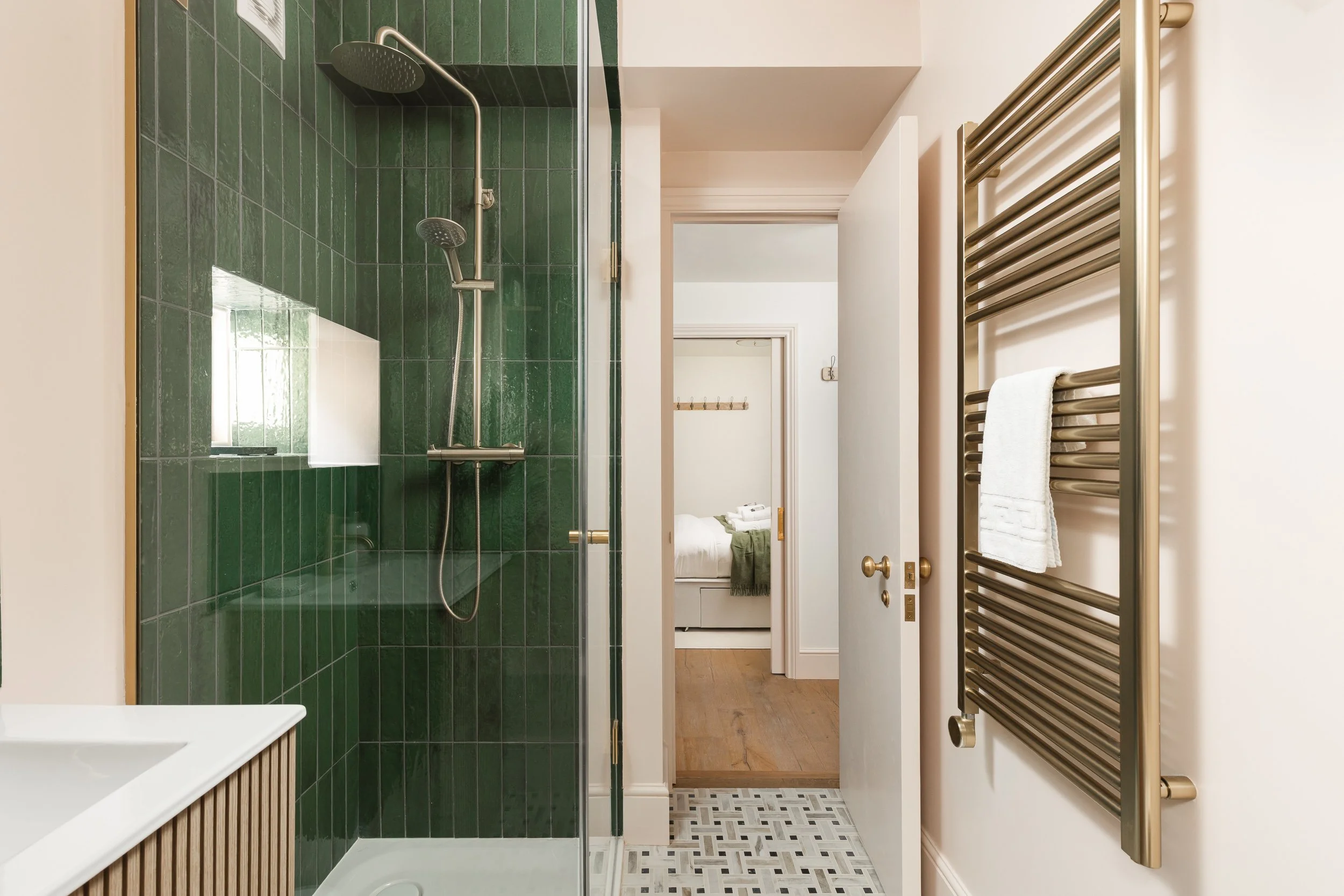 Bathroom with green tiled shower, white walls, and a heated towel rack on the right side with a towel hanging. View into a bedroom through an open door in the background.