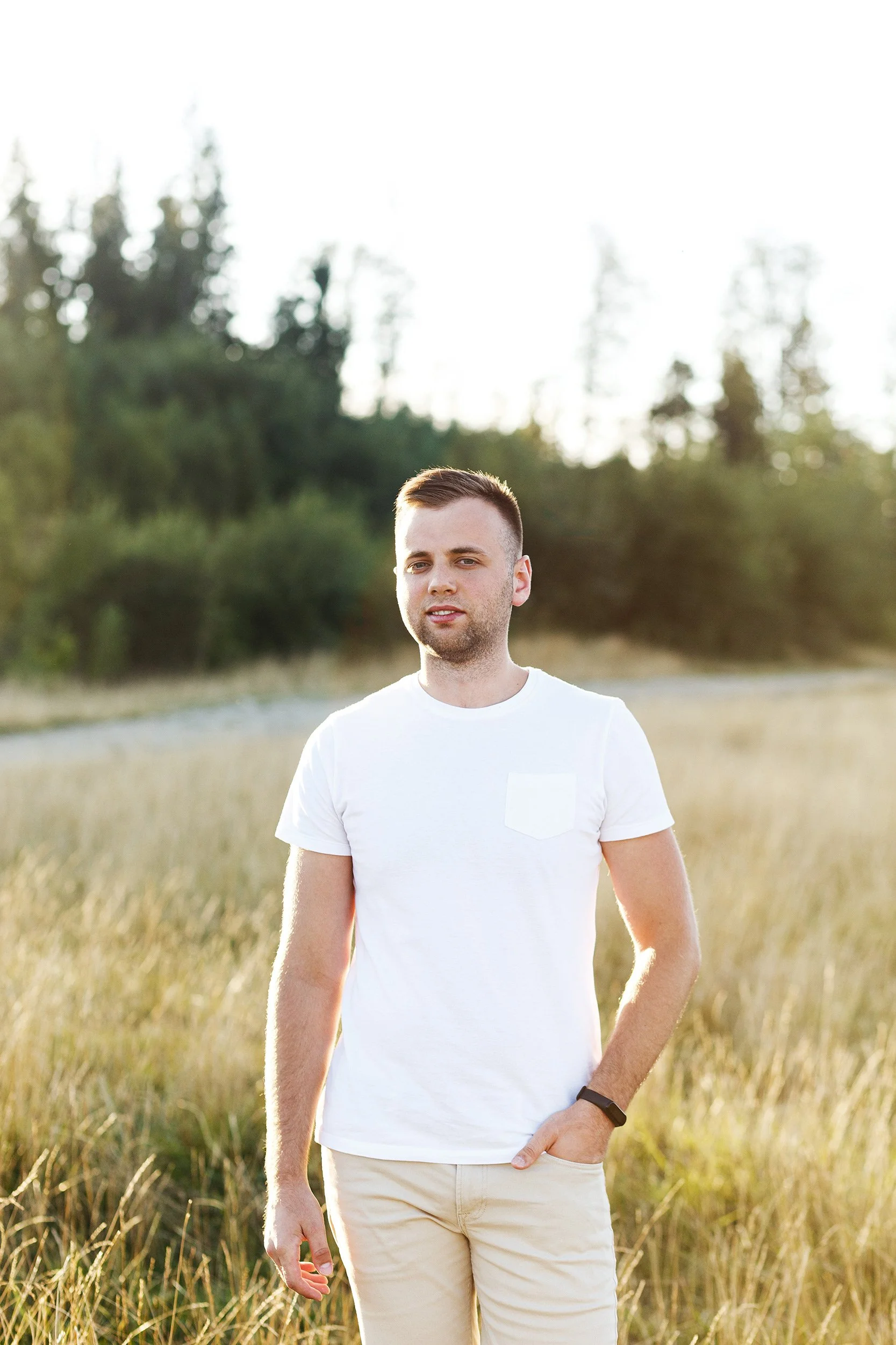 A young man with short brown hair is standing in a field of tall grass, wearing a white t-shirt and beige pants, with a backdrop of trees and a bright, cloudy sky.