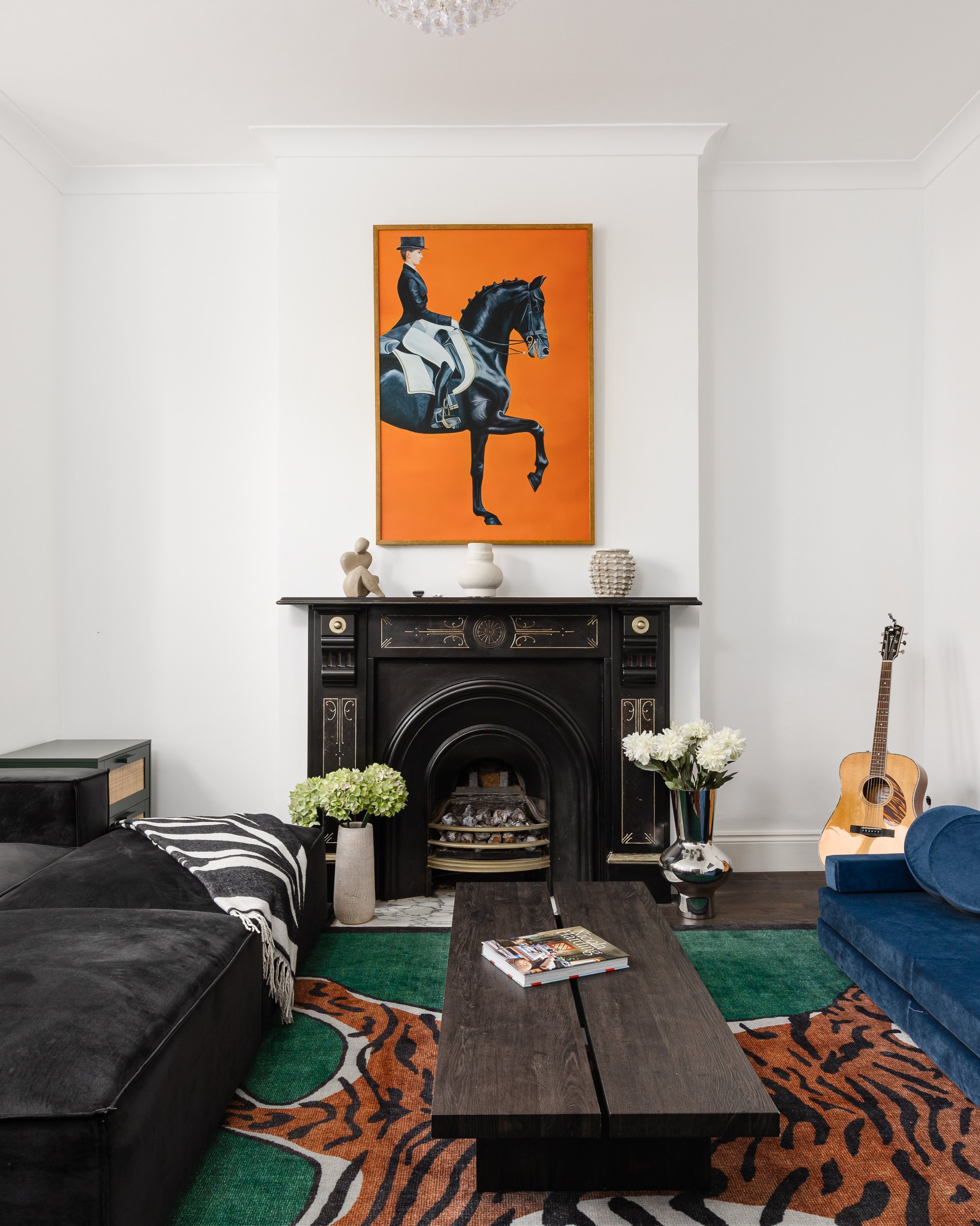 A living room with a black fireplace, artwork of a woman riding a horse on a bright orange background, various vases and sculptures on the mantel, a blue armchair, an acoustic guitar, and a colorful animal print rug.