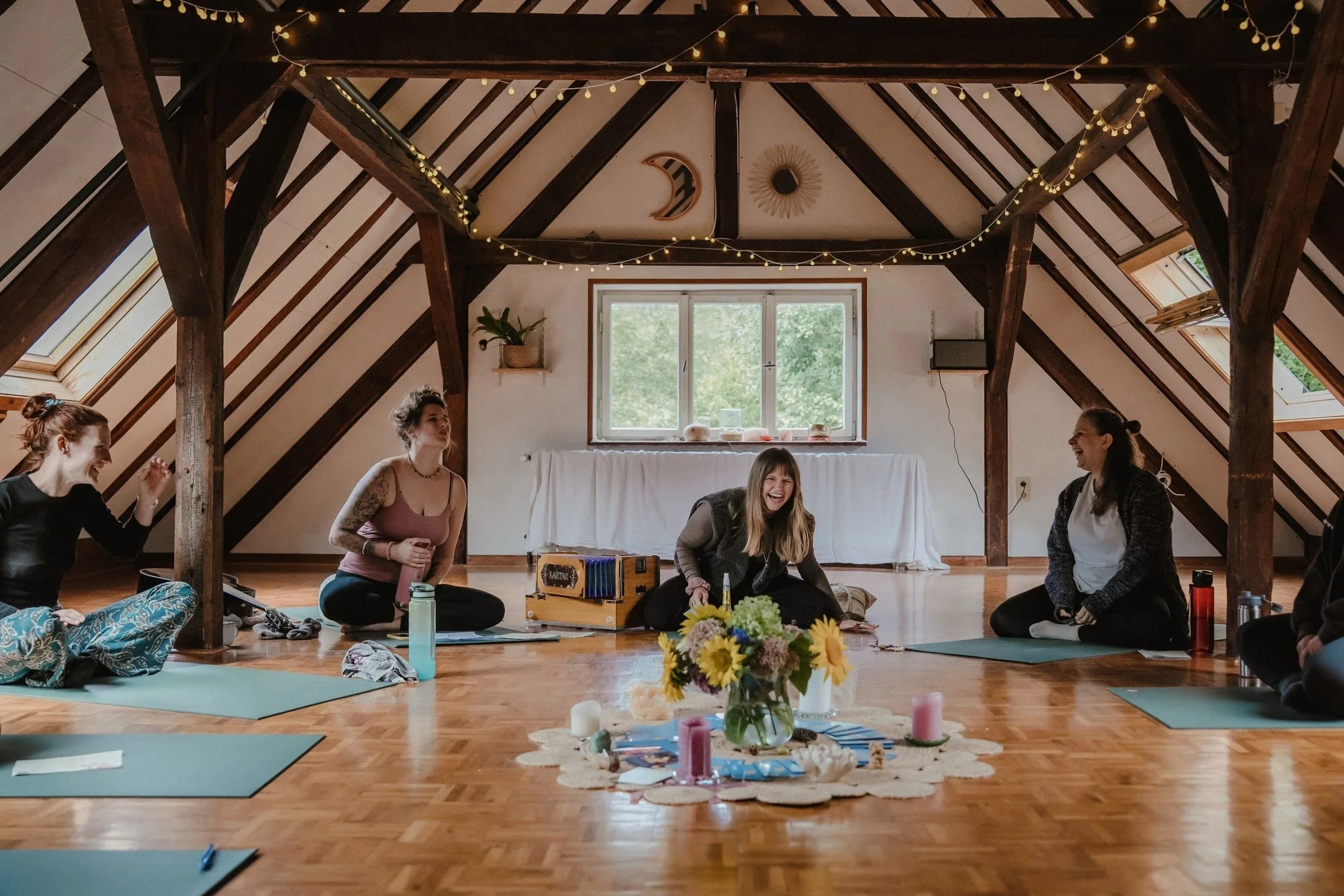 Fünf Frauen sitzen im Kreis auf dem Boden in einem Dachzimmer, lachen und unterhalten sich. Der Raum ist mit Zierlichtern und zwei Wanddekoarten dekoriert und hat eine große Fensterfront mit grüner Natur draußen. Vor ihnen auf dem Boden sind Blumensträuße, Kerzen, Notizblätter und eine Holzbox mit Spielen.