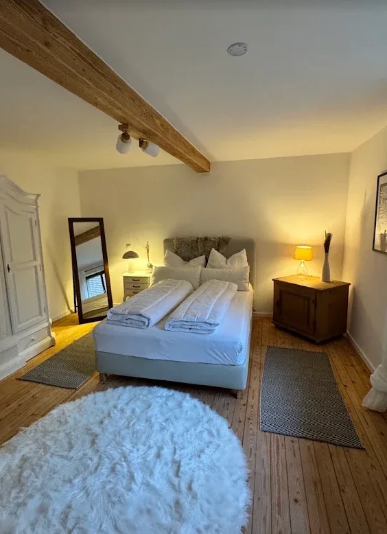 Ein gemütliches Schlafzimmer mit einem Doppelbett, Seitentische mit Lampen, einem großen weißen Kleiderschrank, einem Spiegel, einem Holzfußboden und einem weißen, flauschigen Teppich.