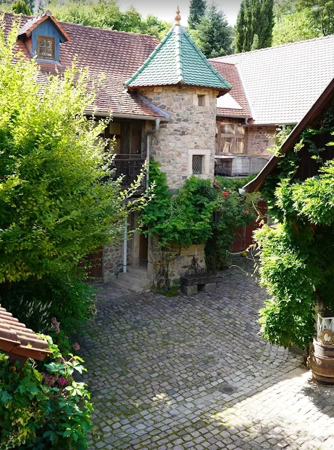 Ein malerischer Innenhof mit ungepflastertem Boden, umgeben von alten Stein- und Holzgebäuden mit Dachbalken und einem kleinen Turm. Viele grüne Bäume und Pflanzen schmücken den Hof, darunter ein großer Baum und Blumen in Töpfen.