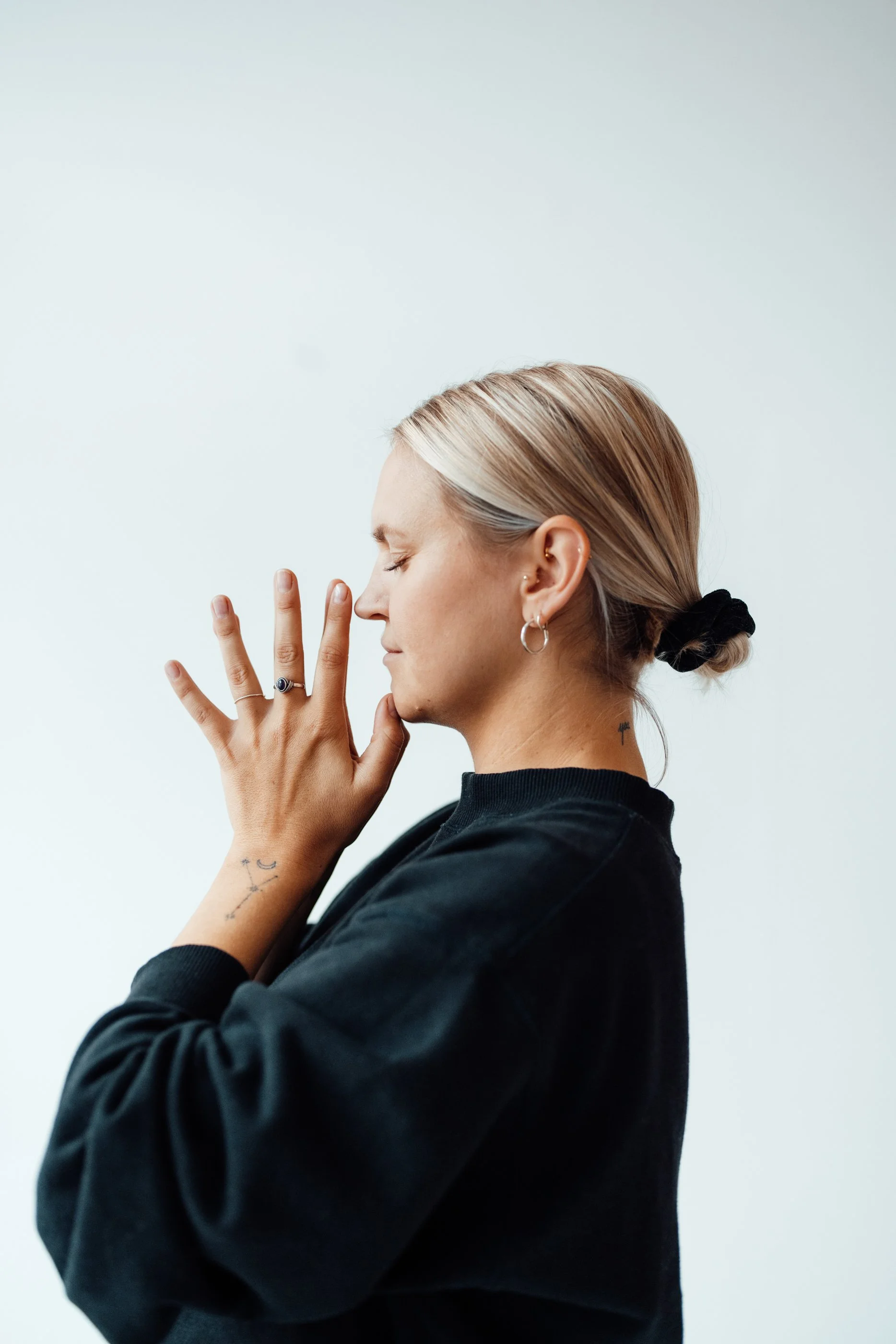Junge Frau mit Tattoos und Ohrringen, die die Augen geschlossen hat und die Fingerspitzen an ihrer Stirn berührt, gegen einen weißen Hintergrund.