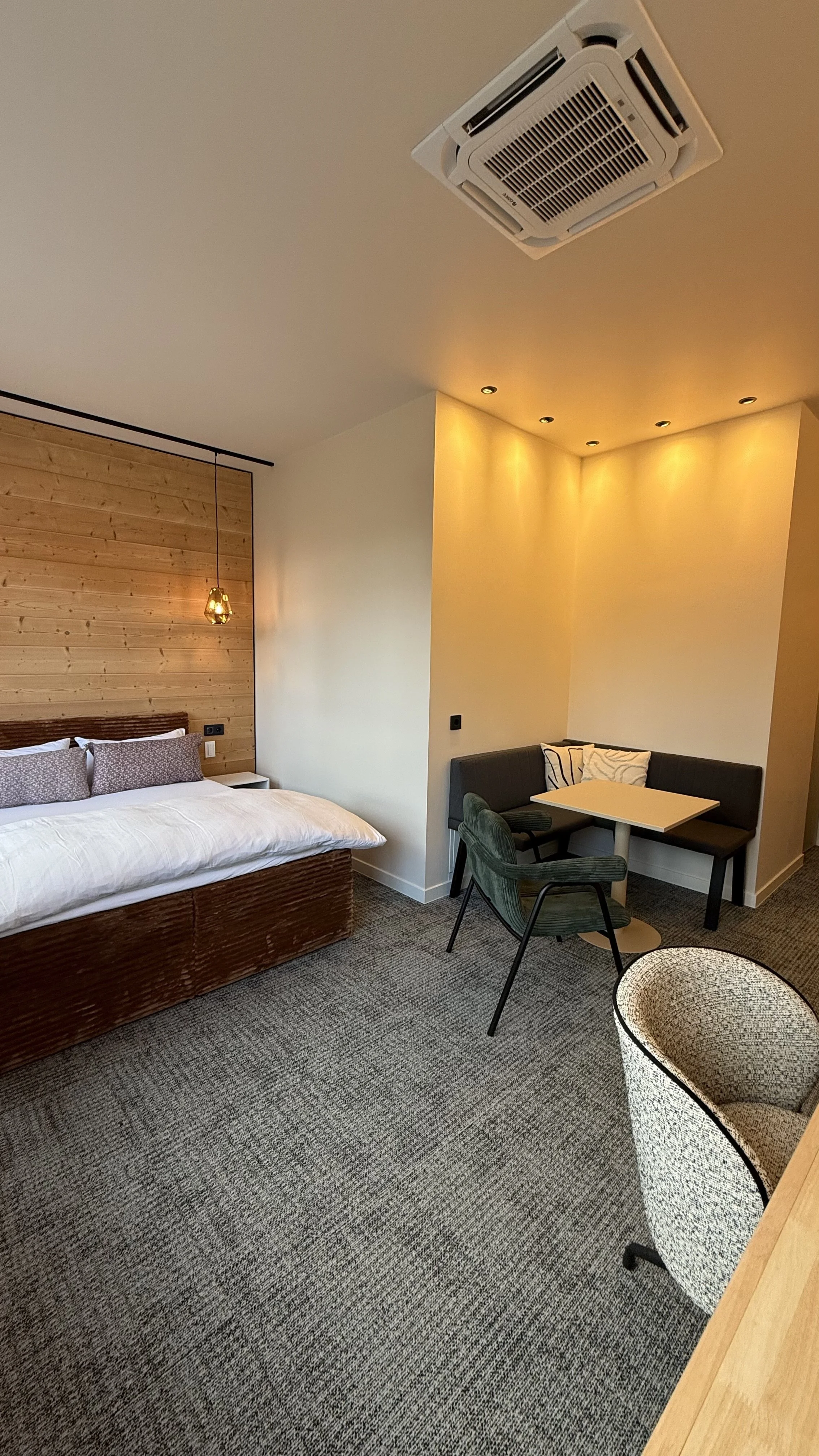 Ein Hotelzimmer mit einem Doppelbett, einer kleinen Sitzecke mit Tisch und Stühlen, eine Wand mit Holzpaneelen und eine moderne Beleuchtung. Es gibt auch eine Klimaanlage an der Decke.