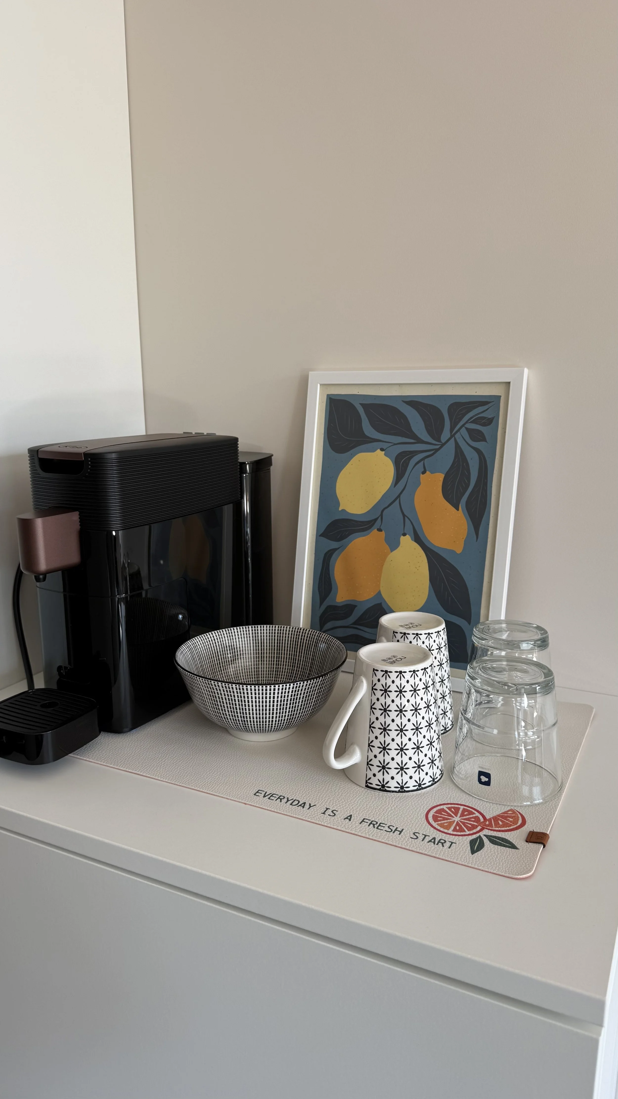 Küchenutensilien auf einer weißen Arbeitsfläche, darunter eine Kaffeemaschine, eine obstförmige Wanddekoration, eine Schüssel, zwei Tee- oder Kaffeetassen, Gläser und ein Tischset mit dem Spruch "Everyday is a fresh start".