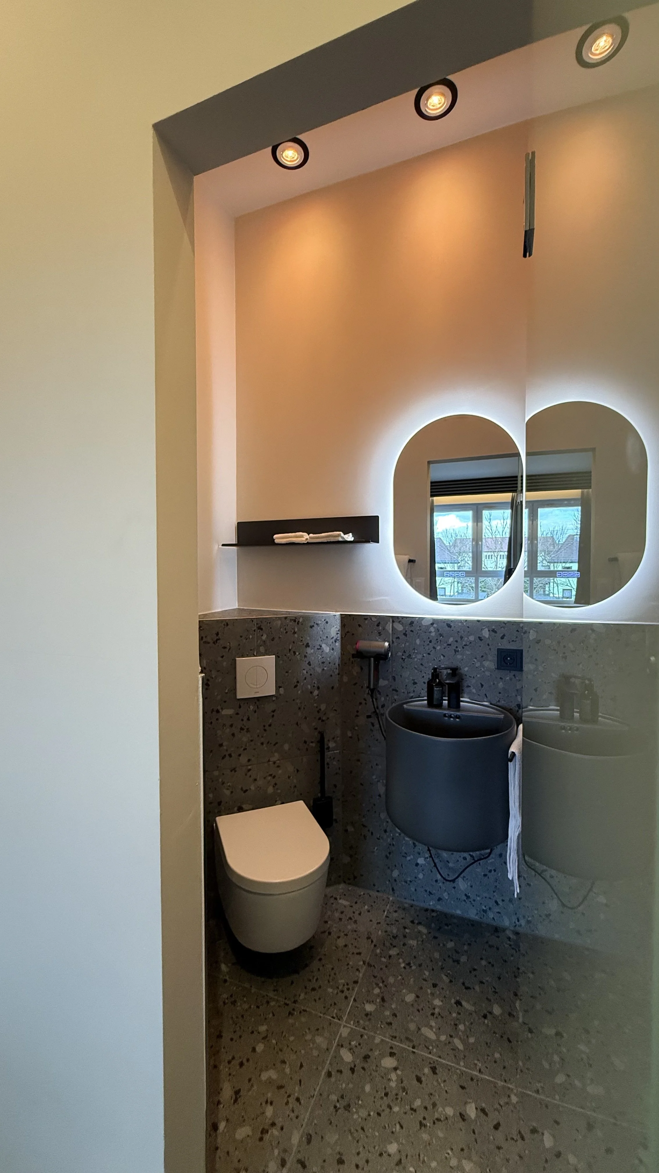 Moderner Badezimmerraum mit Spiegel, Waschbecken, WC, dunkler Fliesenspiegel, Fenster und Deckenleuchten.