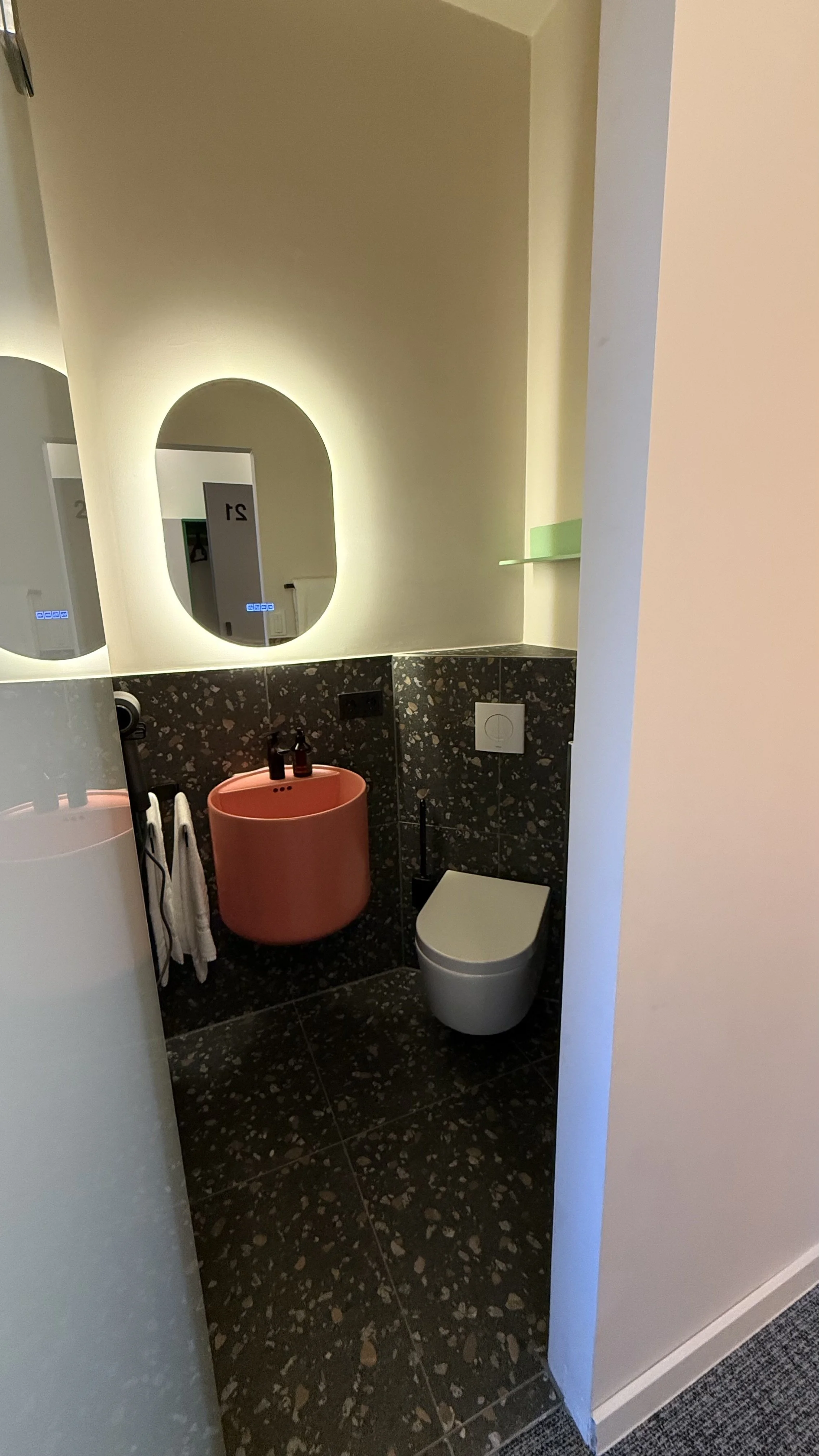 Ein modernes Badezimmer mit einer Wandtoilette, einem schwarzen Fliesenboden, einem runden Waschbecken in Rostfarbe, einem runden Wandspiegel mit Licht und persönlichen Pflegeprodukten auf dem Waschbecken.