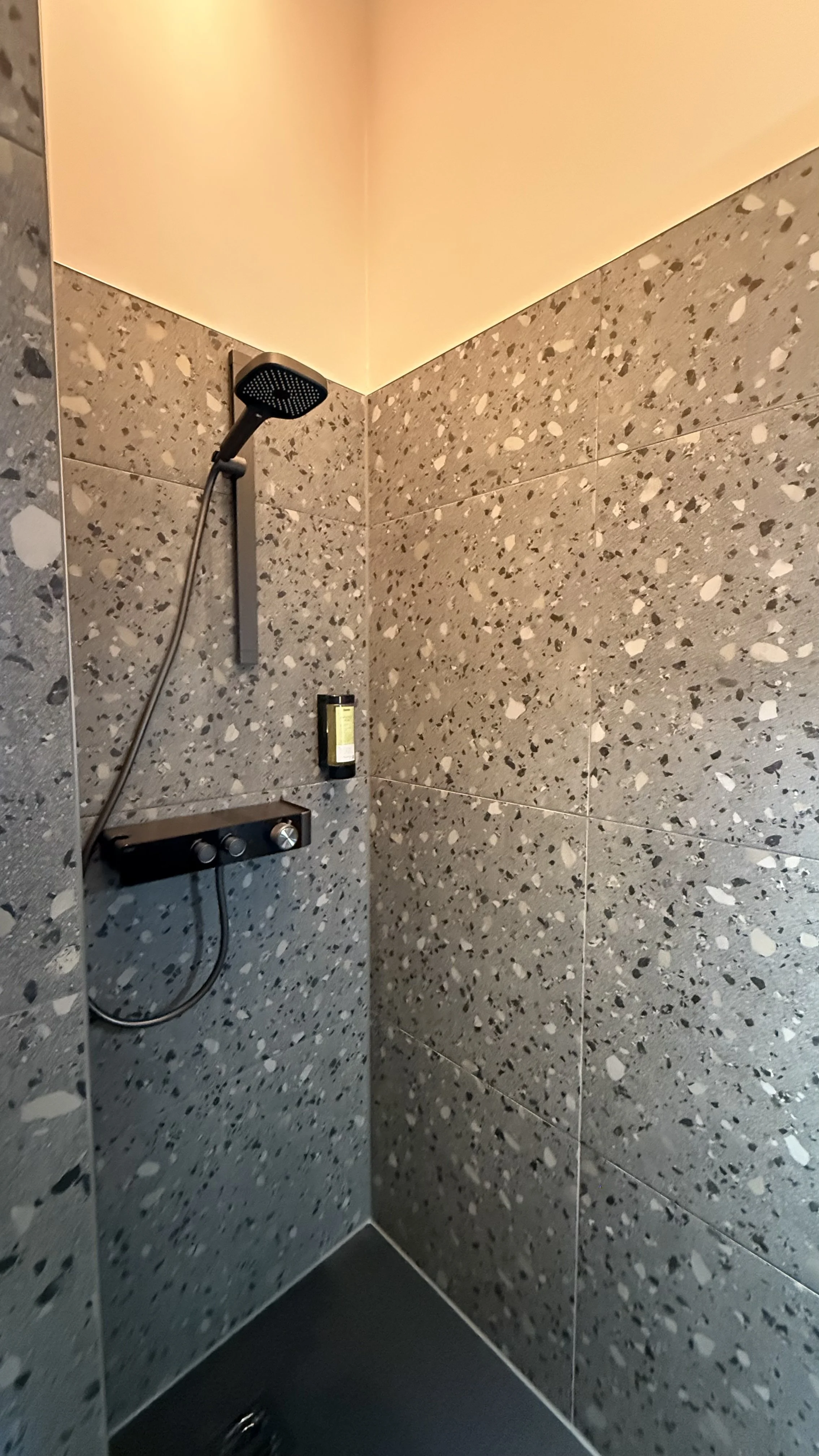 Dusche mit dunklem Duschkopf, Armatur und Pflegeprodukt in einer Wandnische, Fliesen mit grauem Marmor-Design.