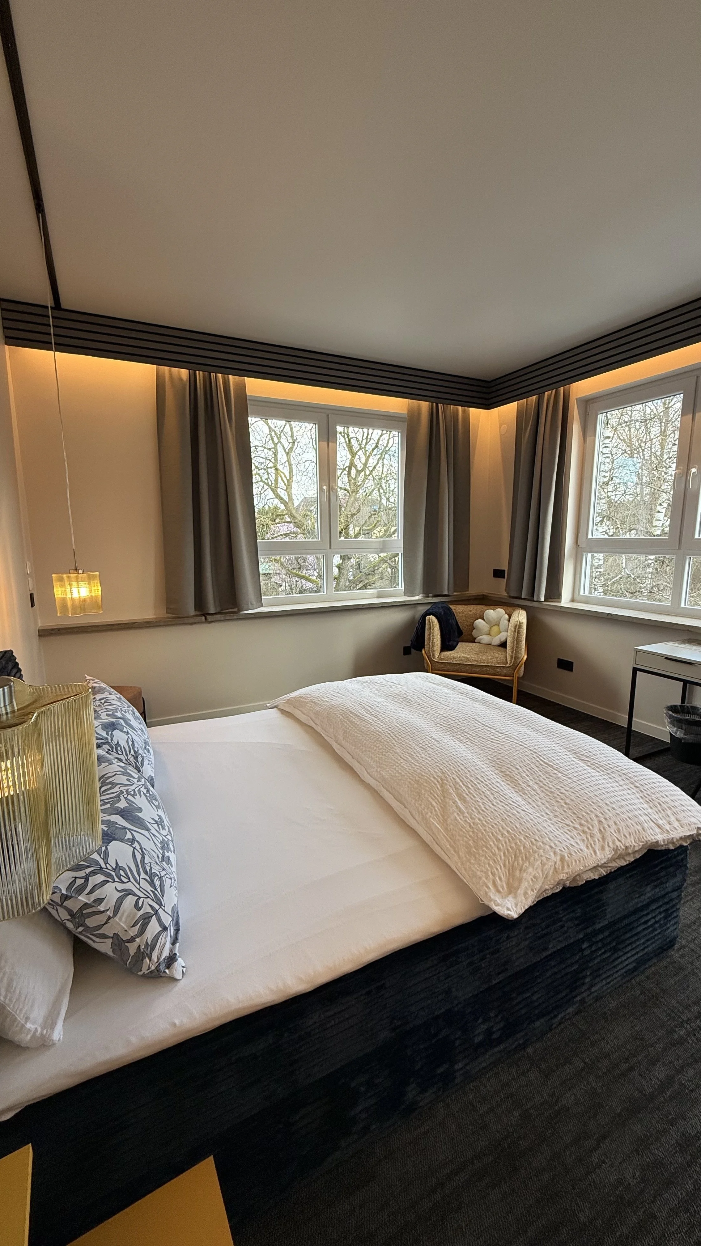 Ein gemütliches Schlafzimmer mit großen Fenstern, einem Bett mit weißen und blauen Bettwäsche, einem Nachttisch mit einer Lampe, einem Sessel mit Kissen und einer kleinen weißen Kommode. Die Fenster bieten einen Blick auf Bäume im Freien.