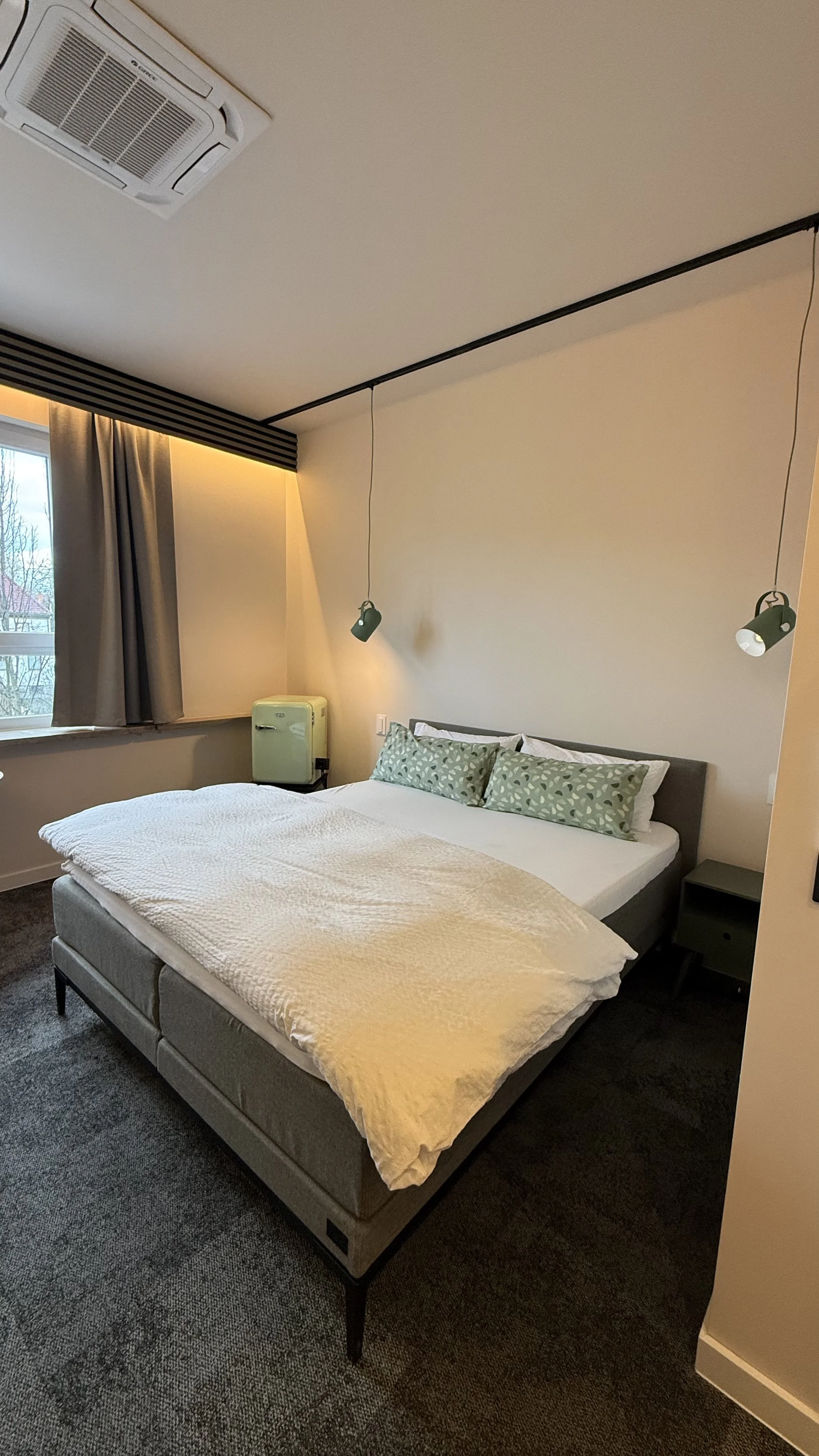 Ein modernes Schlafzimmer mit einem großen Bett, zwei grüngemusterten Kissen, seitlichen Nachttischen, dunklen Vorhängen, einem Fenster, einer Deckenlufteinheit und Wandleuchten.