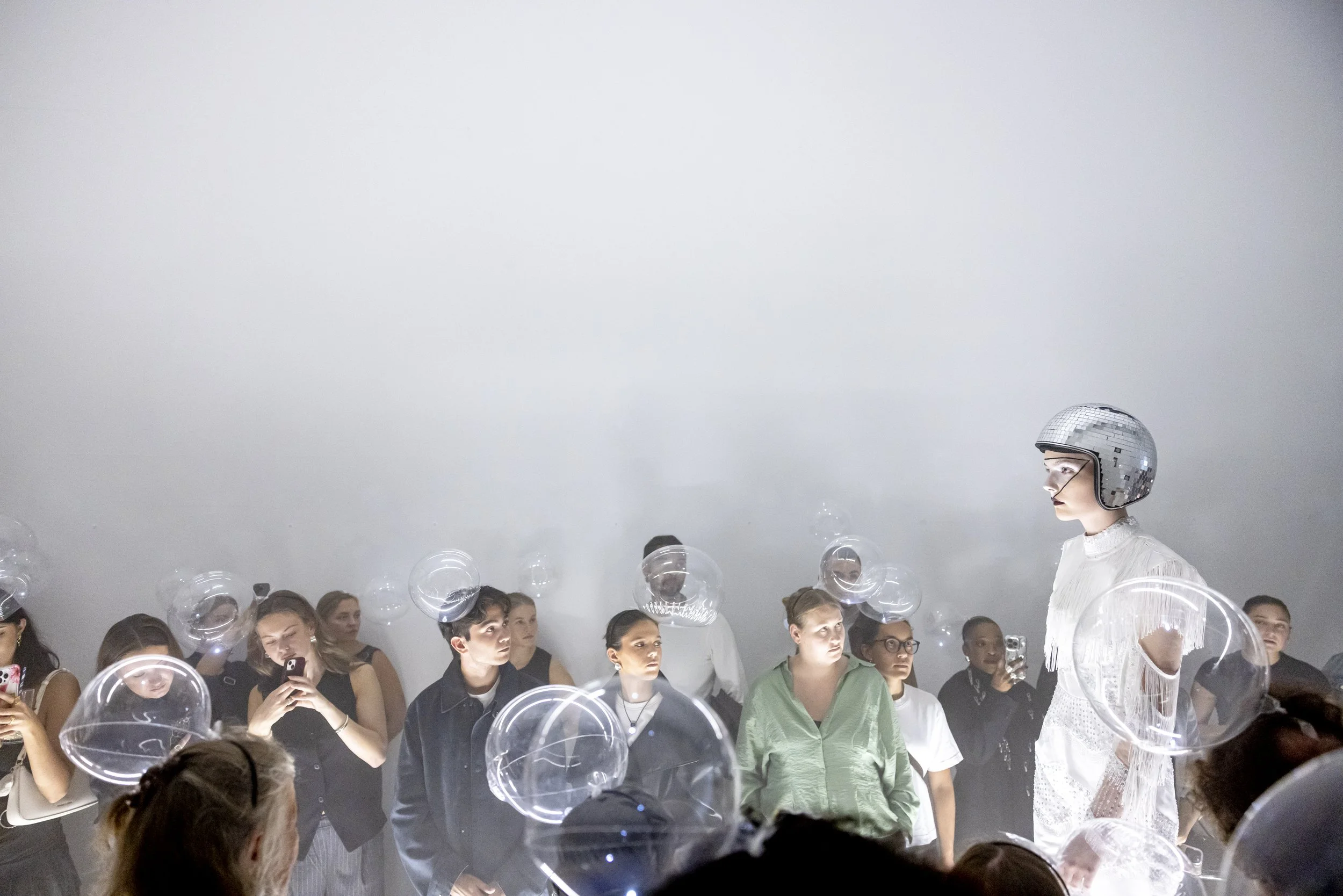 A woman in a white dress and silver helmet performs for an audience, with bubbles around her, as audience members look and take pictures.