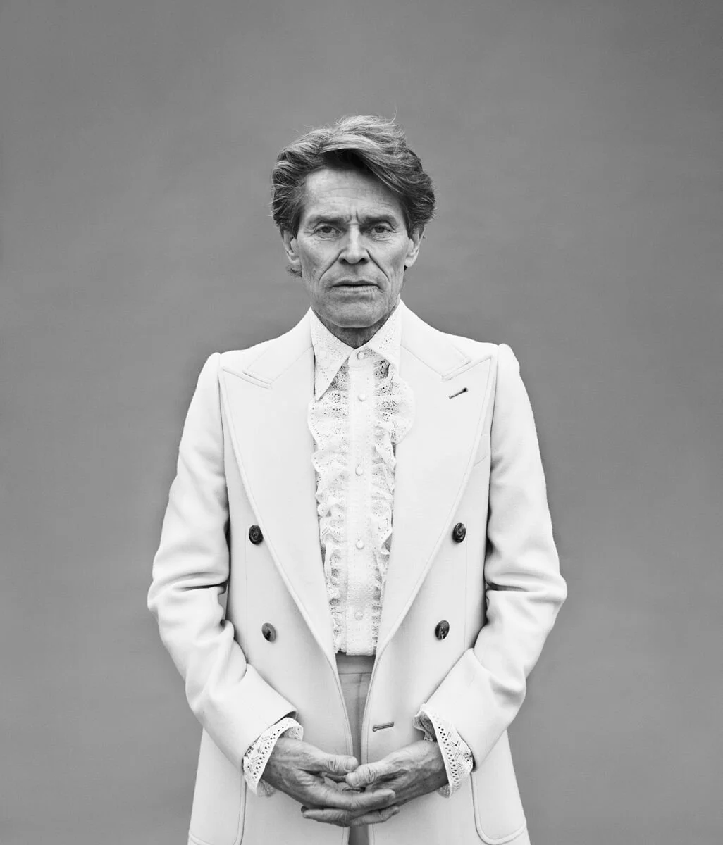 Black and white portrait of an older man with styled hair, wearing a Victorian-style lace shirt and a modern light-colored double-breasted suit jacket, standing against a plain background.