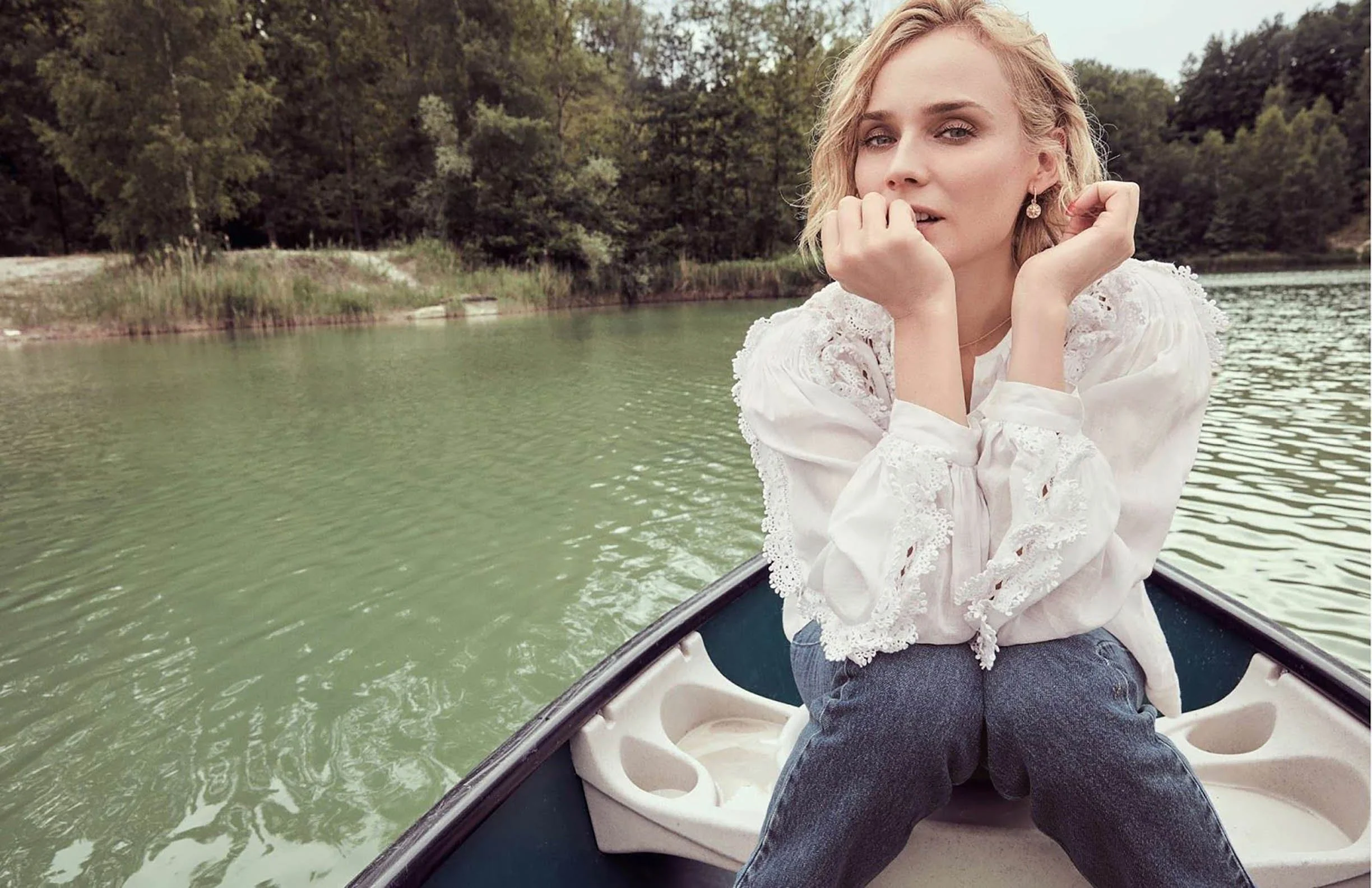 A woman with blonde curly hair and earrings is sitting in a boat on a calm green lake, wearing a white lace blouse and dark jeans, with a wooded shoreline in the background.