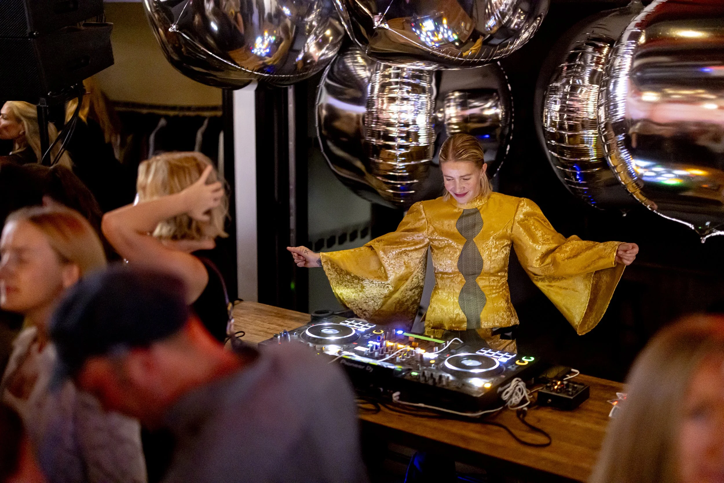 A woman in a shiny yellow dress is DJing at a party, standing behind a DJ setup with large silver balloons in the background, while people gather around enjoying the event.