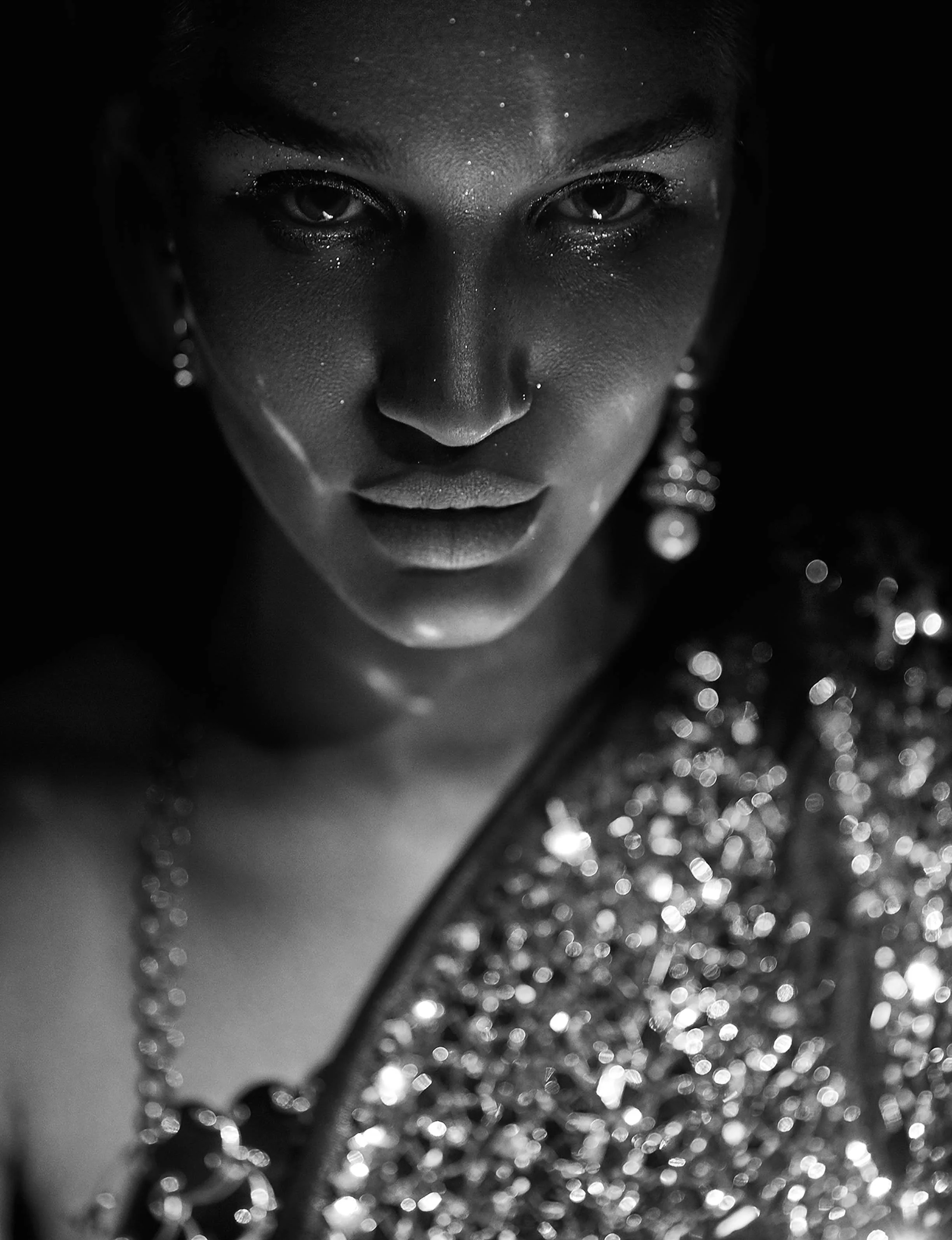 Close-up black and white photo of a woman with dark skin, shiny makeup, jewelry, and a sequined garment.